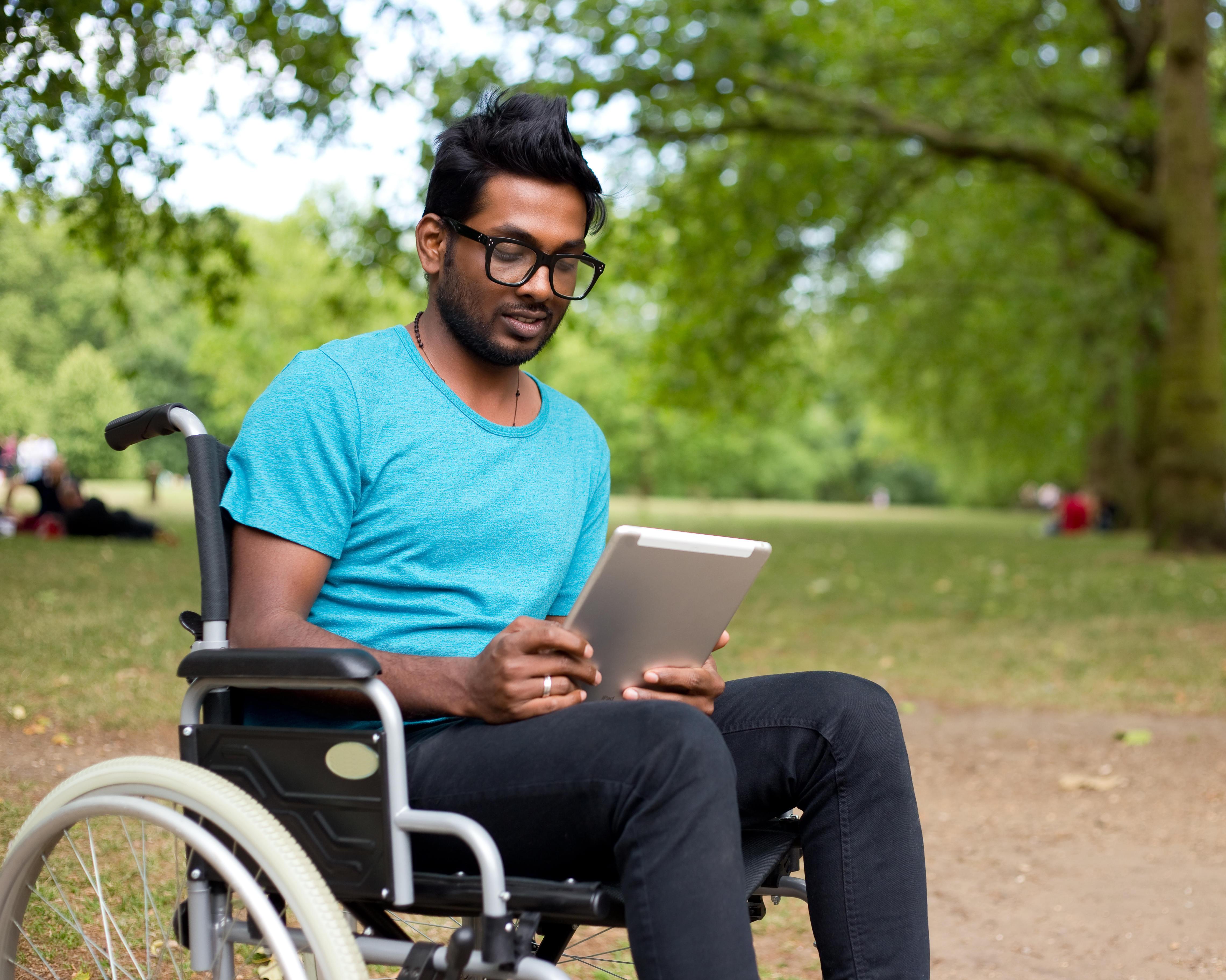 Man using a wheelchair reads a tablet outdoors in a green park.
