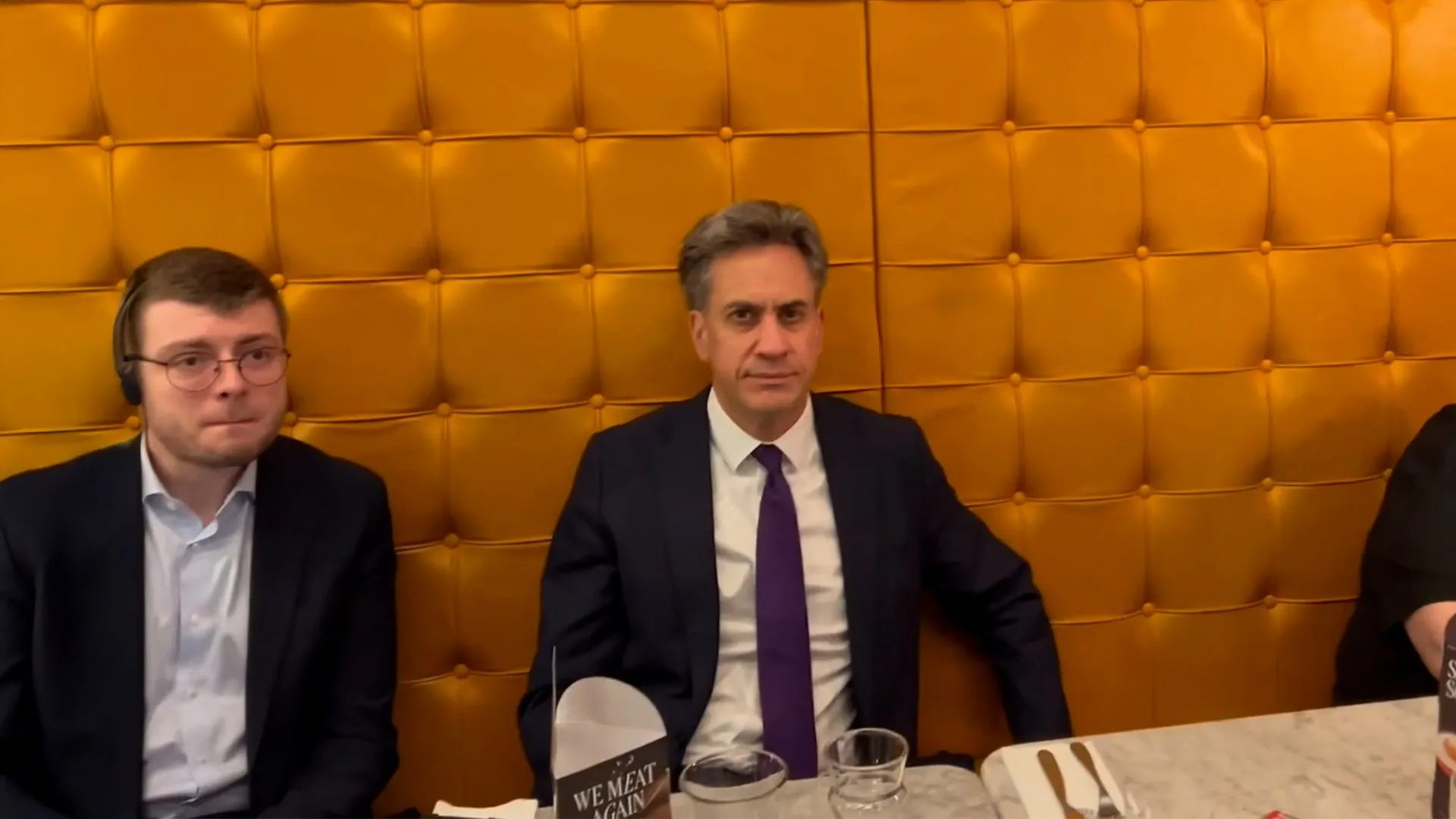 Two suited men sit at a restaurant table against an orange padded wall, with a third person partly visible.