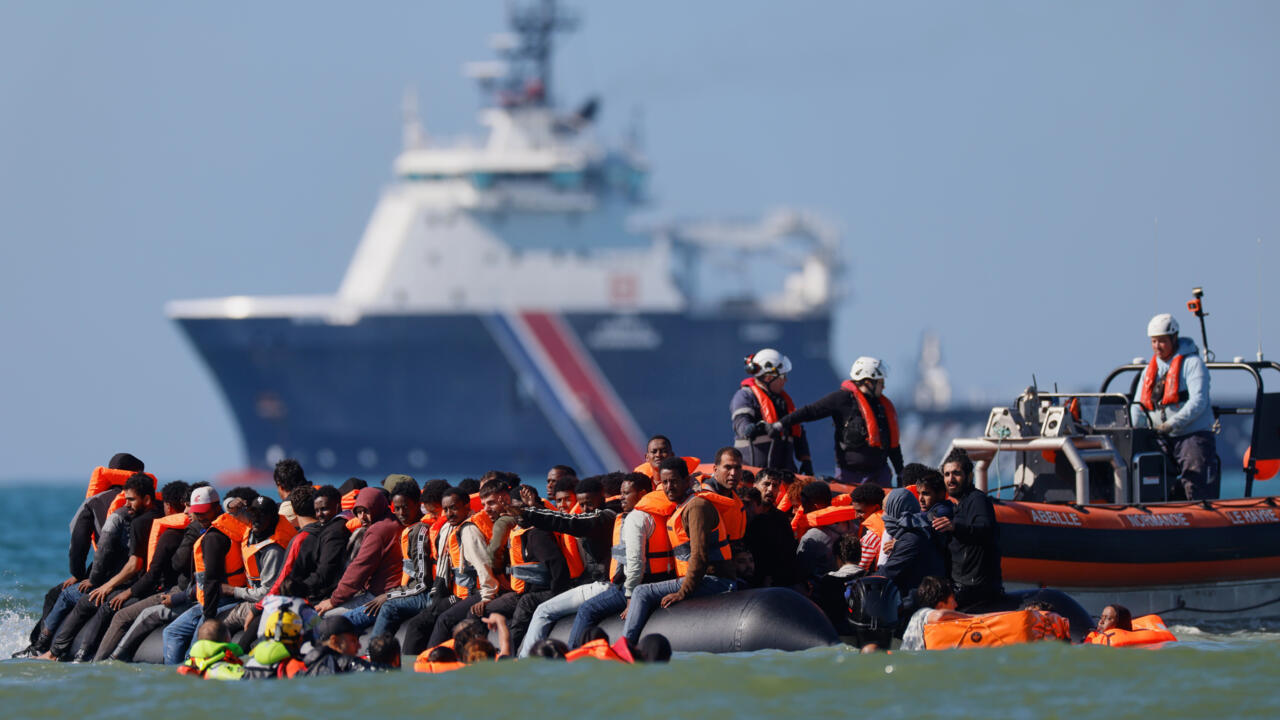 People wearing orange life jackets sit aboard an inflatable boat beside a French rescue vessel and patrol ship.