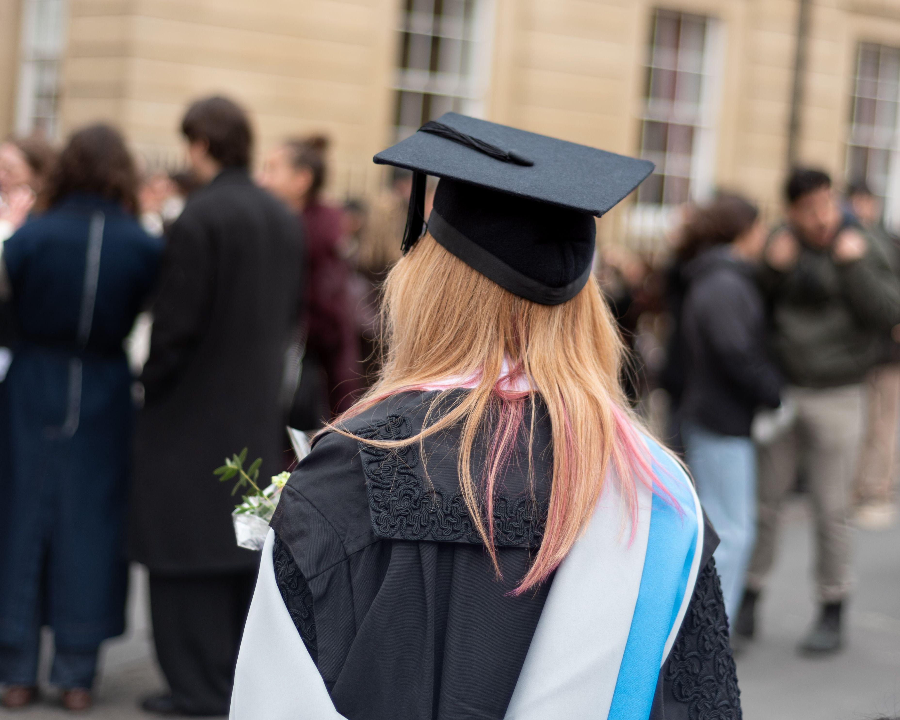 Graduate in cap and gown walking among blurred people outside a university building