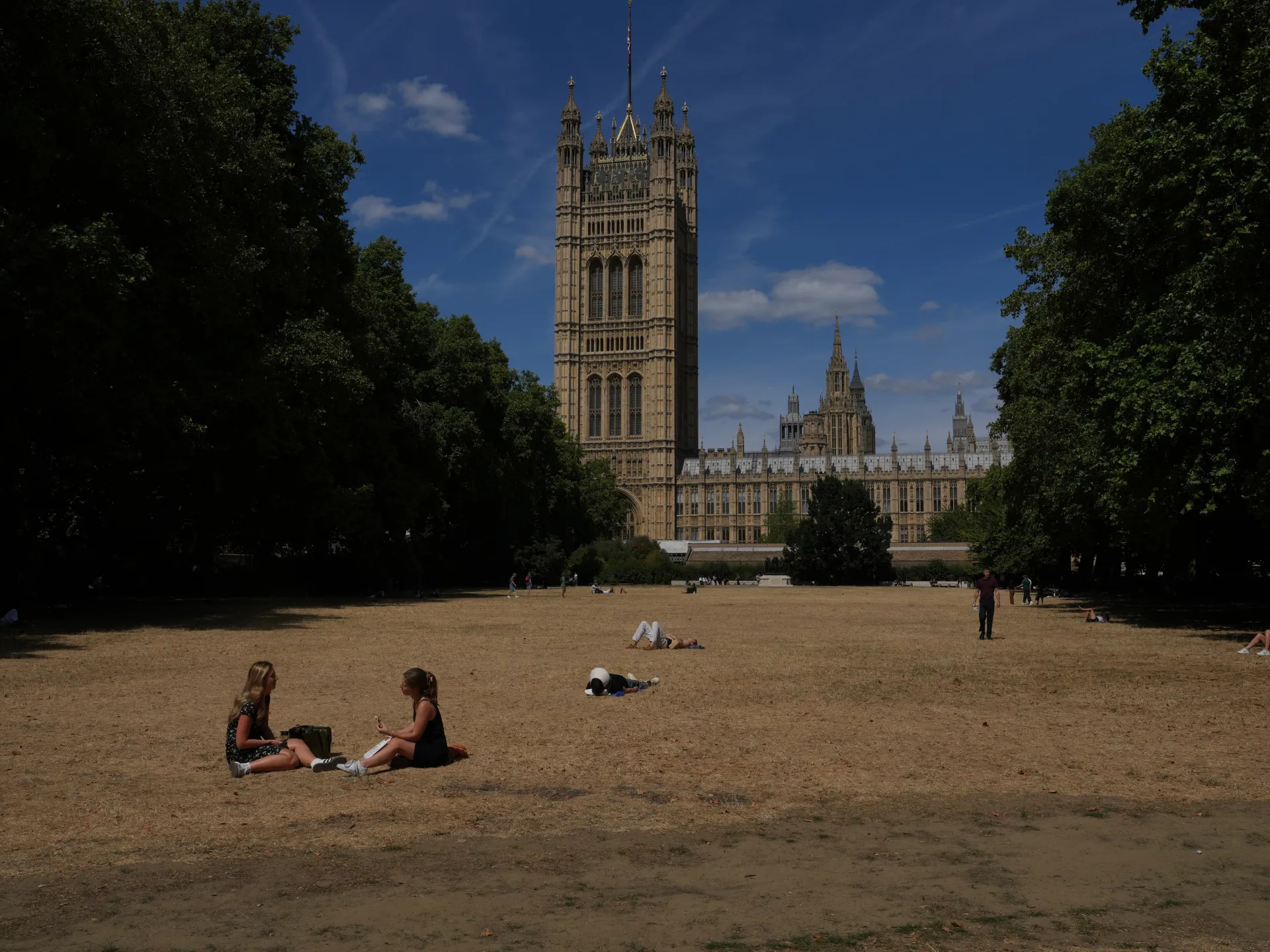 People sit and relax on a dry lawn with the UK Parliament buildings visible behind trees.