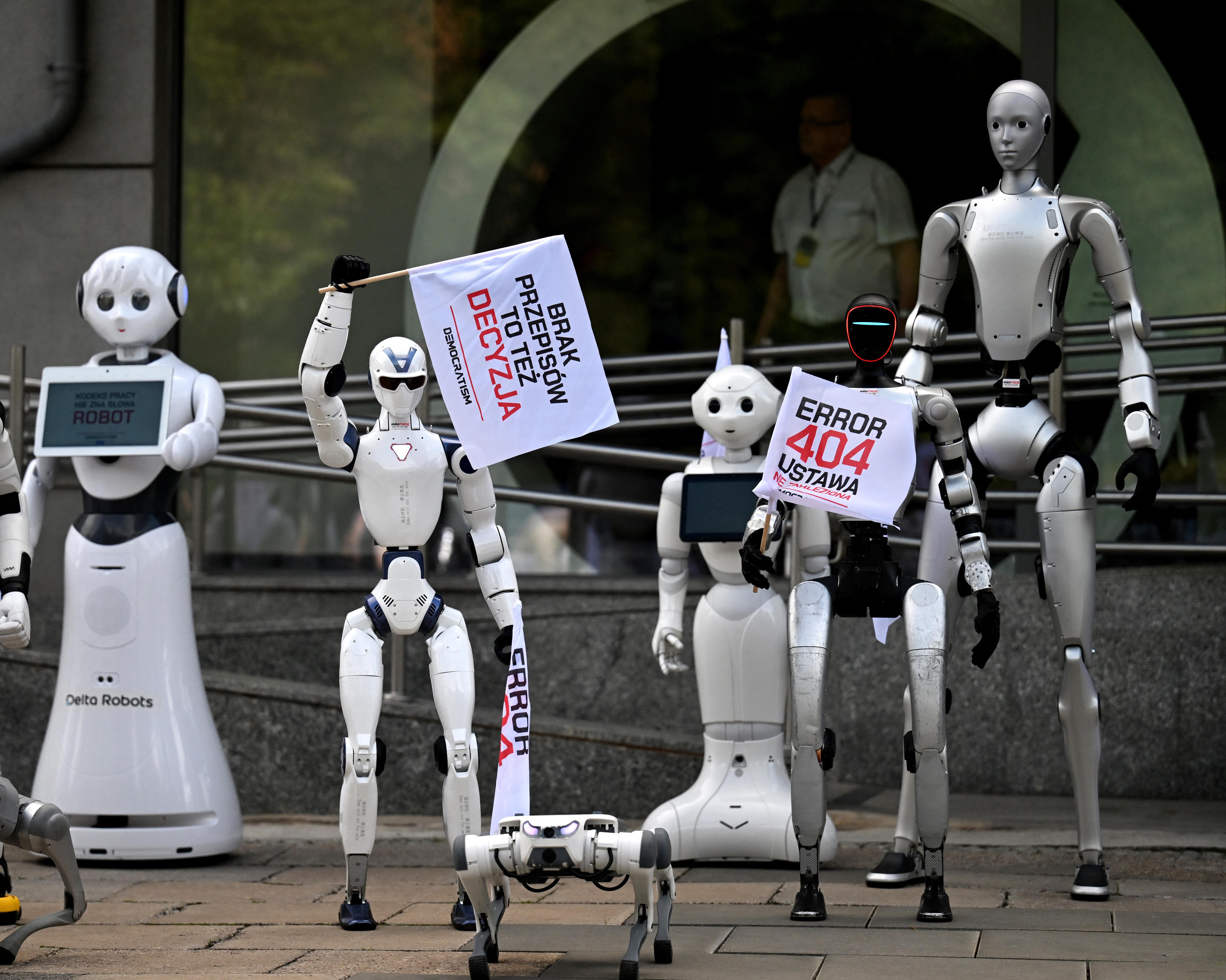 Humanoid robots hold protest placards outside a building, including messages about democracy and AI.