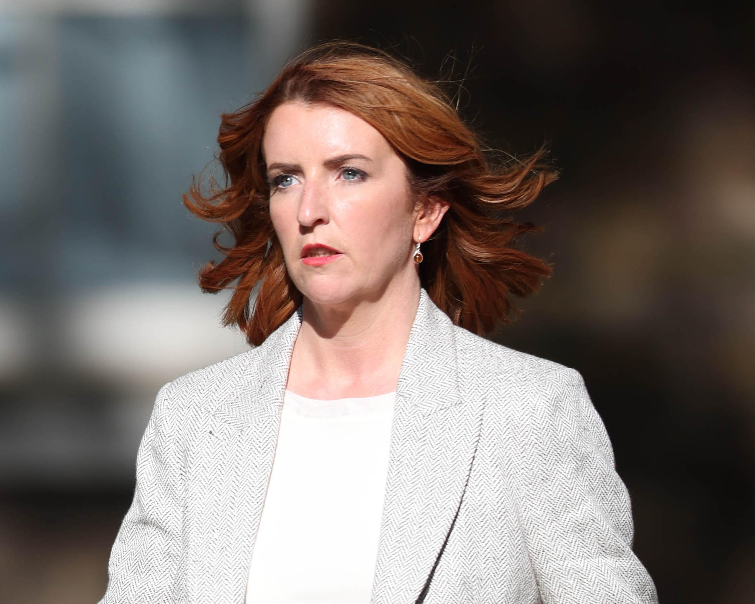 Red-haired woman wearing a light patterned blazer looks ahead against a blurred dark background