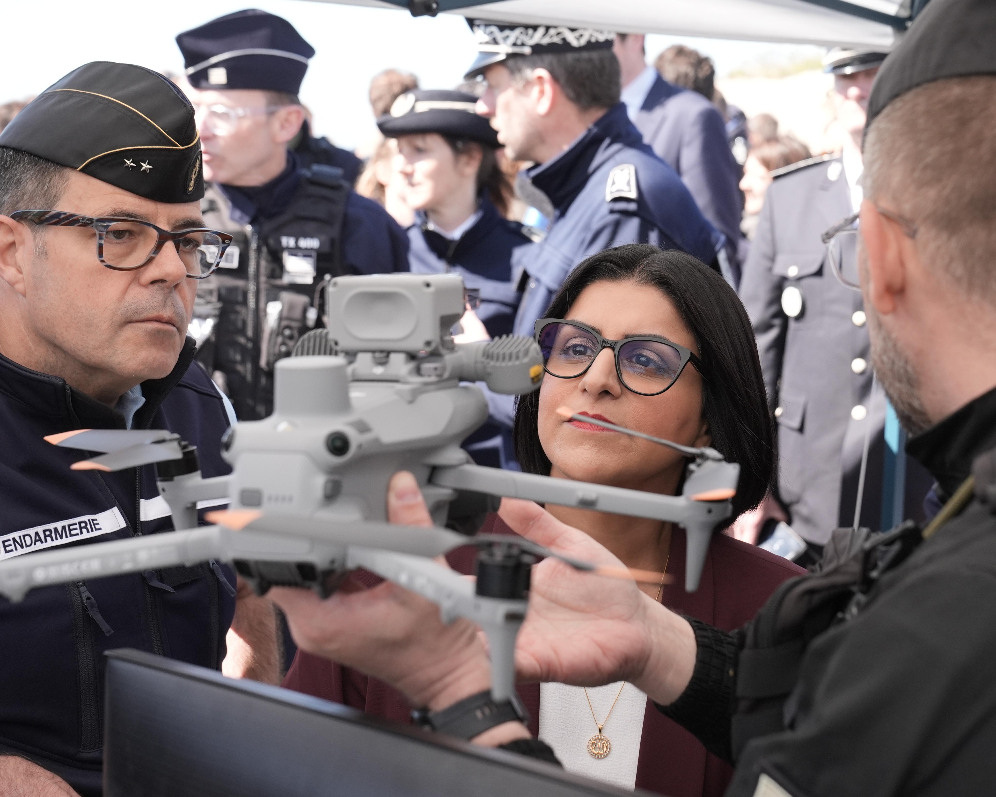 Officials and a woman examine a drone amid a crowded outdoor gathering.