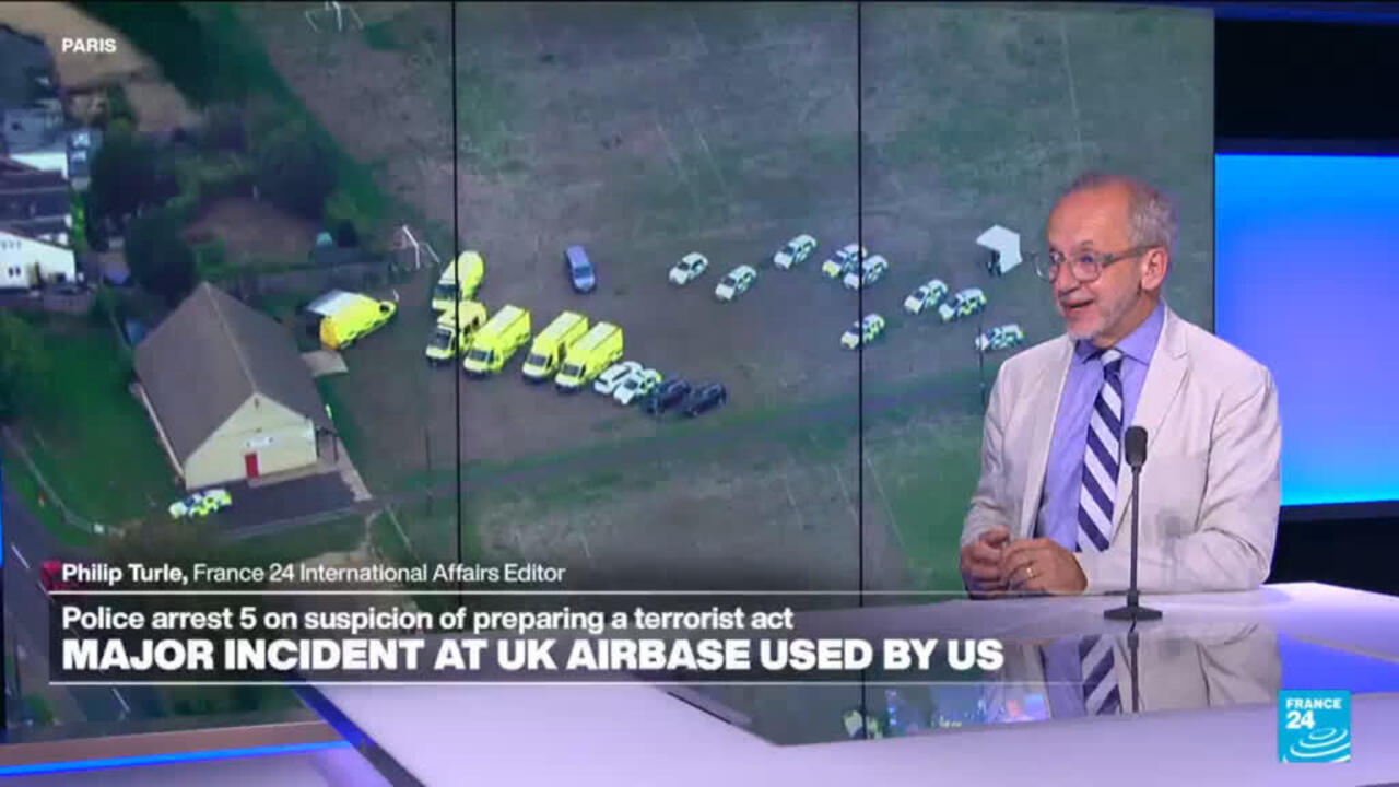 News presenter speaks beside aerial footage showing emergency vehicles gathered near a large rural building.