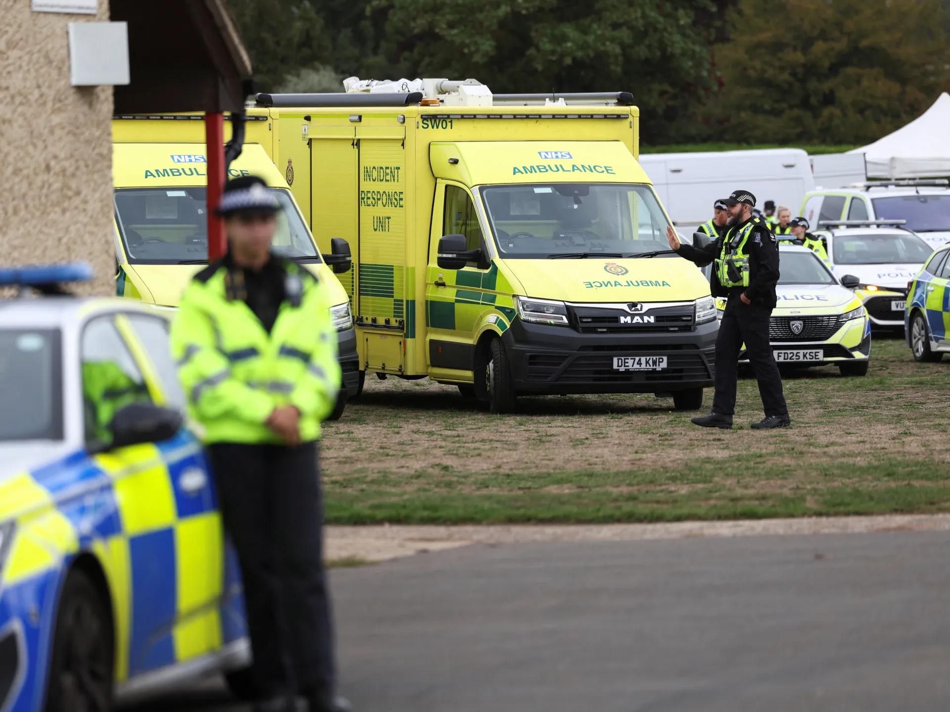 Yellow ambulances and police vehicles gather as uniformed officers stand beside them on a grassy area.