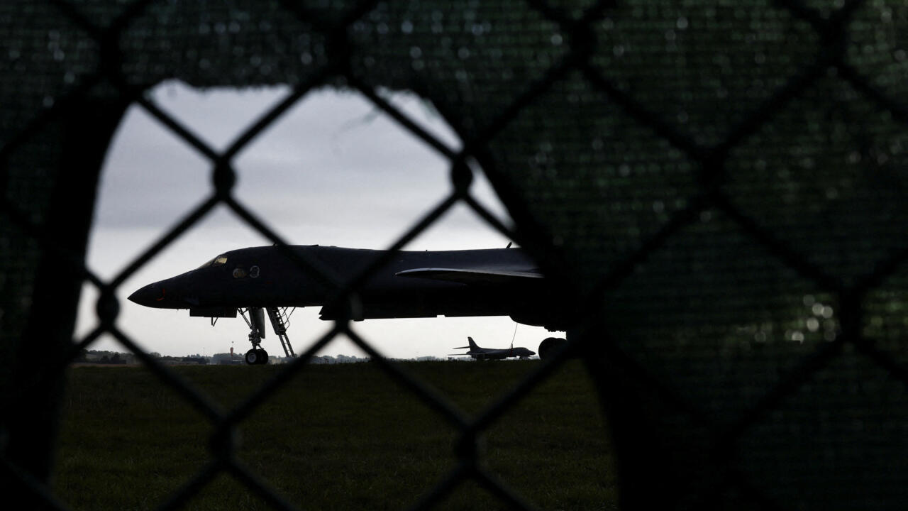 A dark military aircraft sits on a runway behind a chain-link fence, with another plane in the distance.