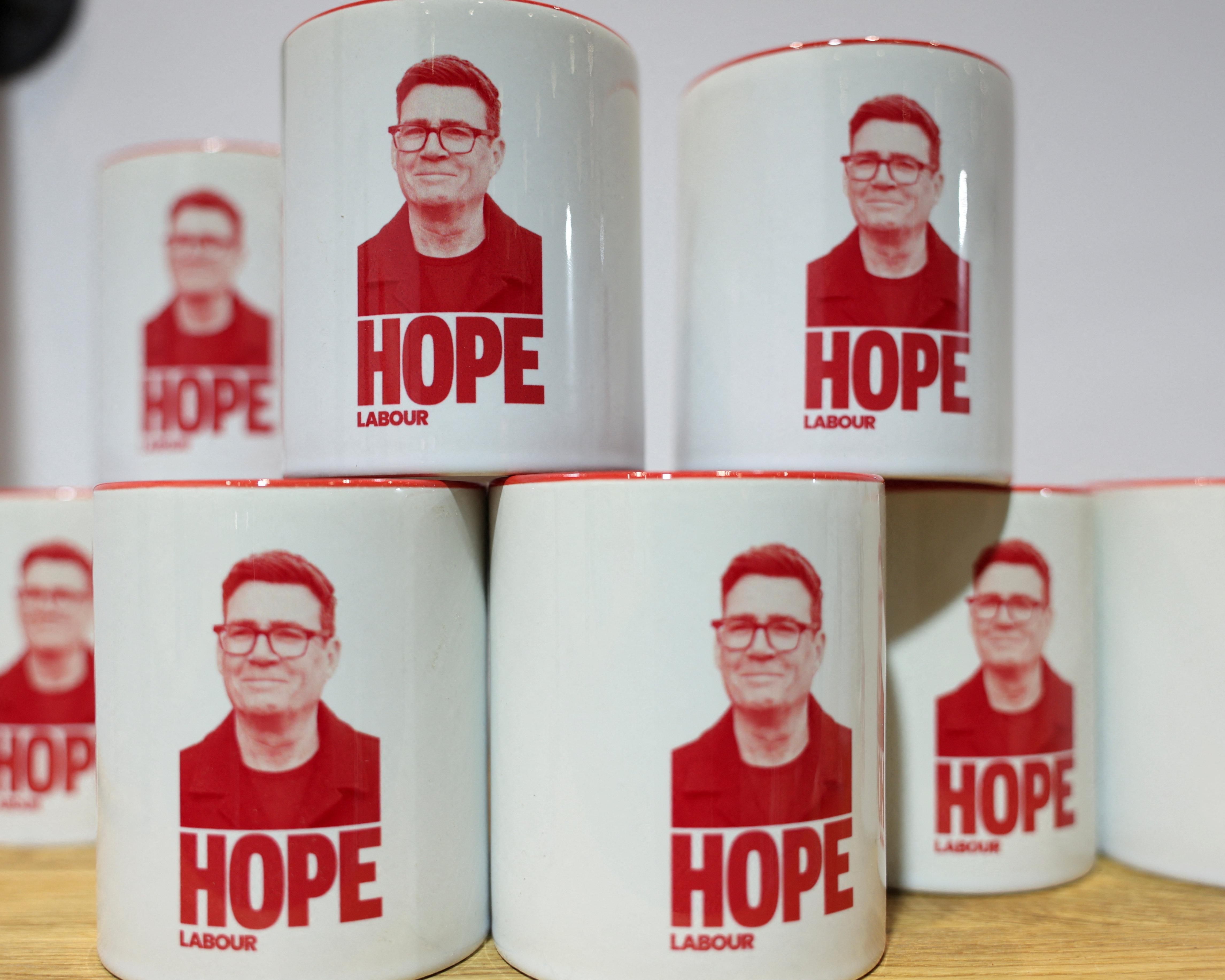 White mugs printed with a red portrait and the words “HOPE LABOUR” stacked on a wooden surface.