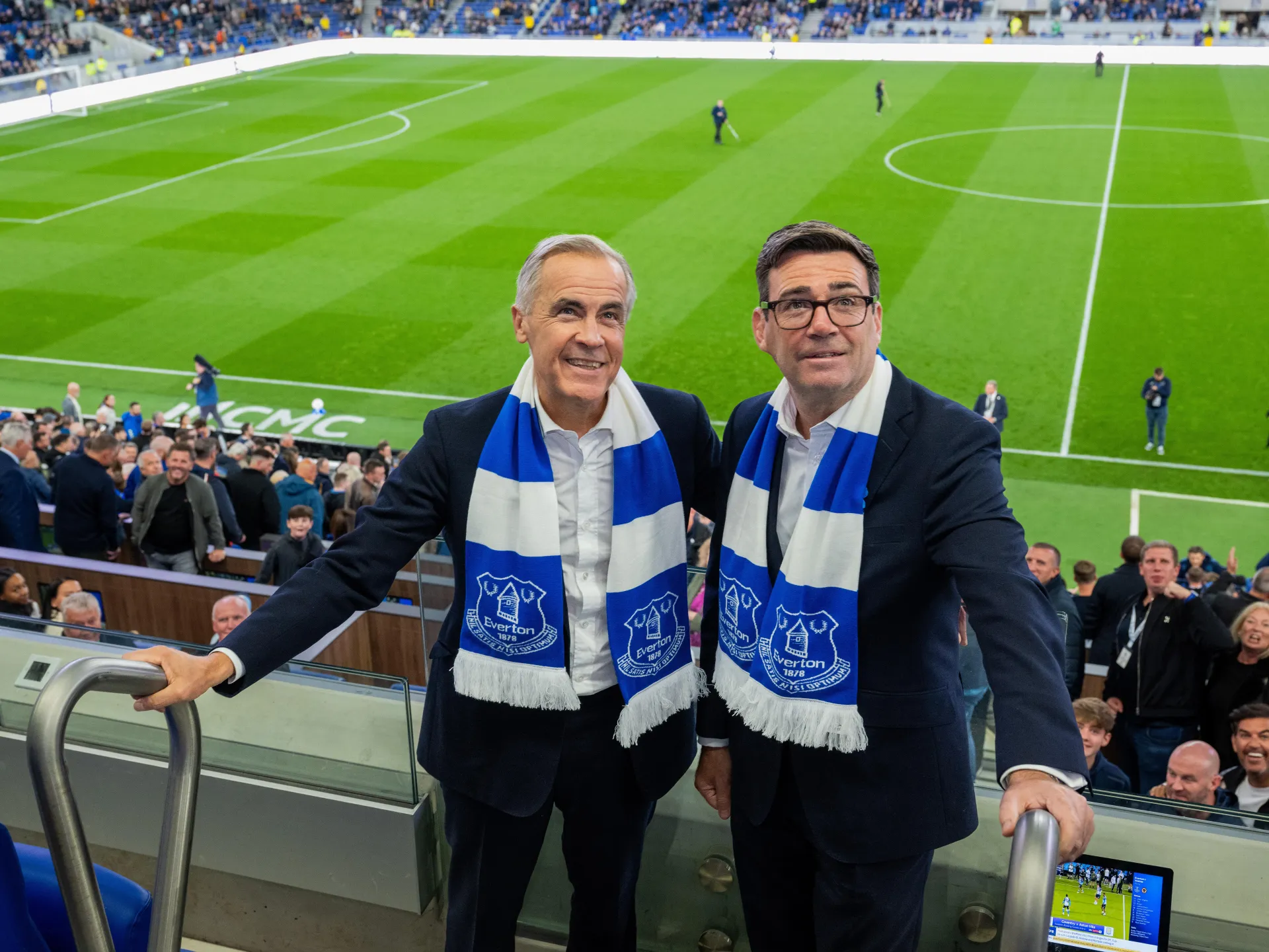 Two suited men wearing blue-and-white Everton scarves pose in stadium seating beside a football pitch.