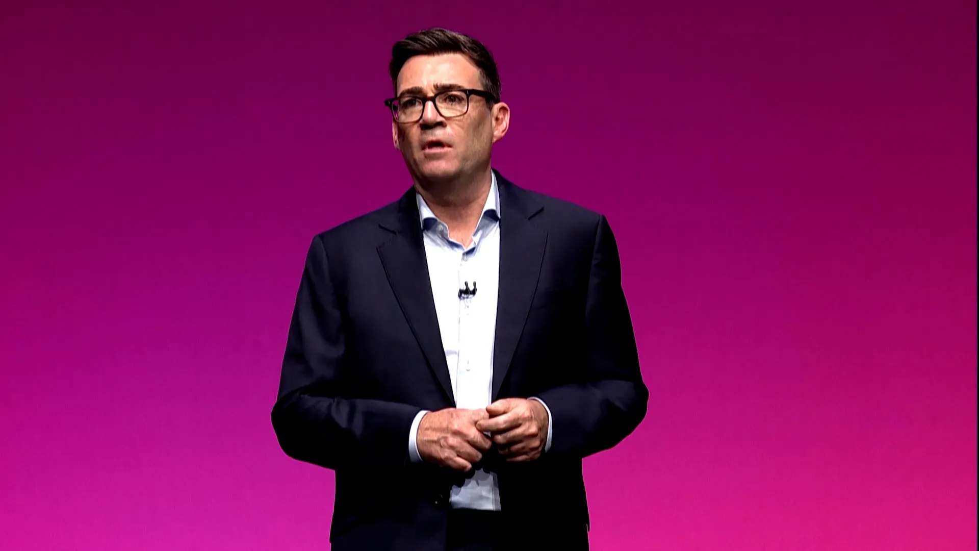 A bespectacled man in a dark suit speaks onstage against a magenta backdrop.