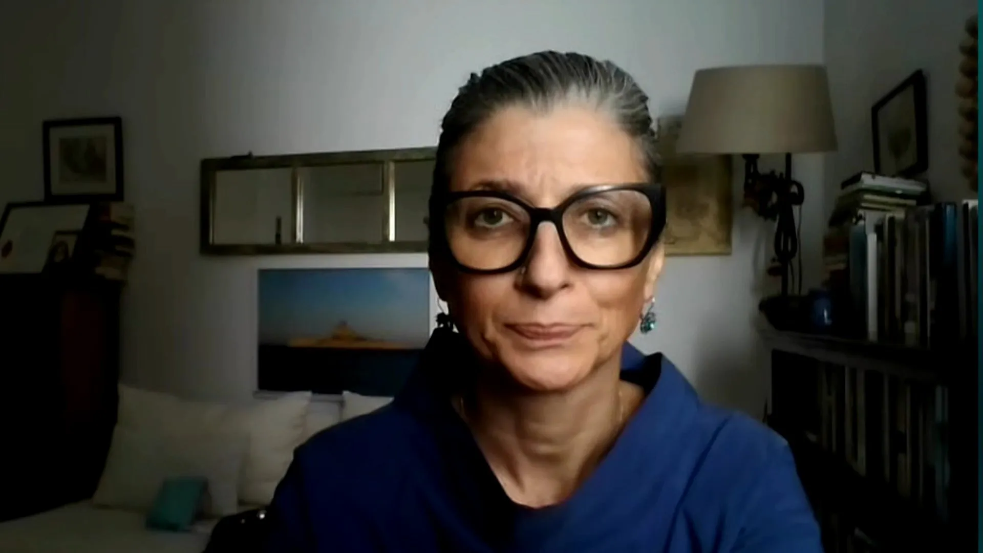 Woman wearing black glasses and a blue top speaks during a video call indoors.