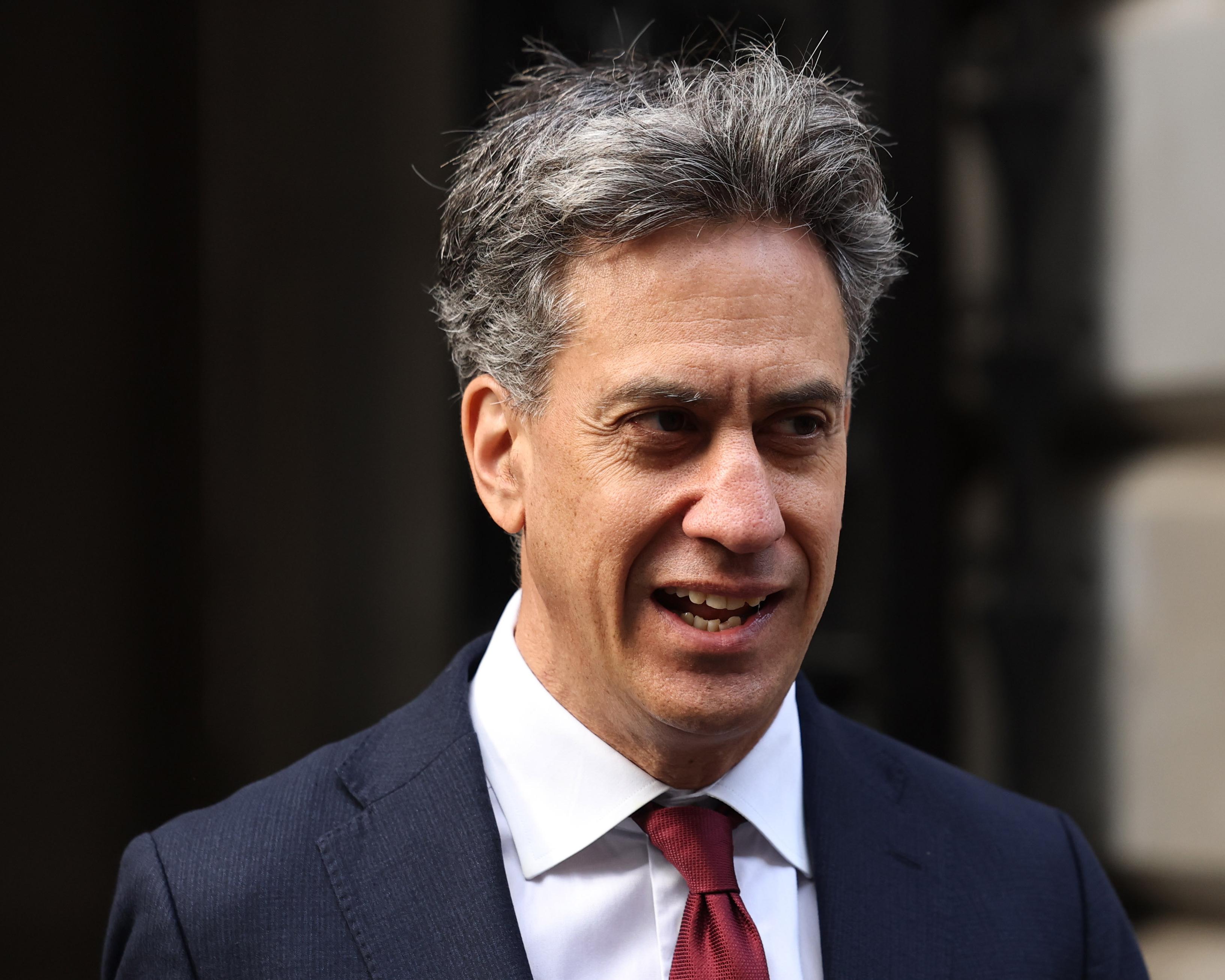 Gray-haired man in navy suit and red tie speaking outdoors near a dark building