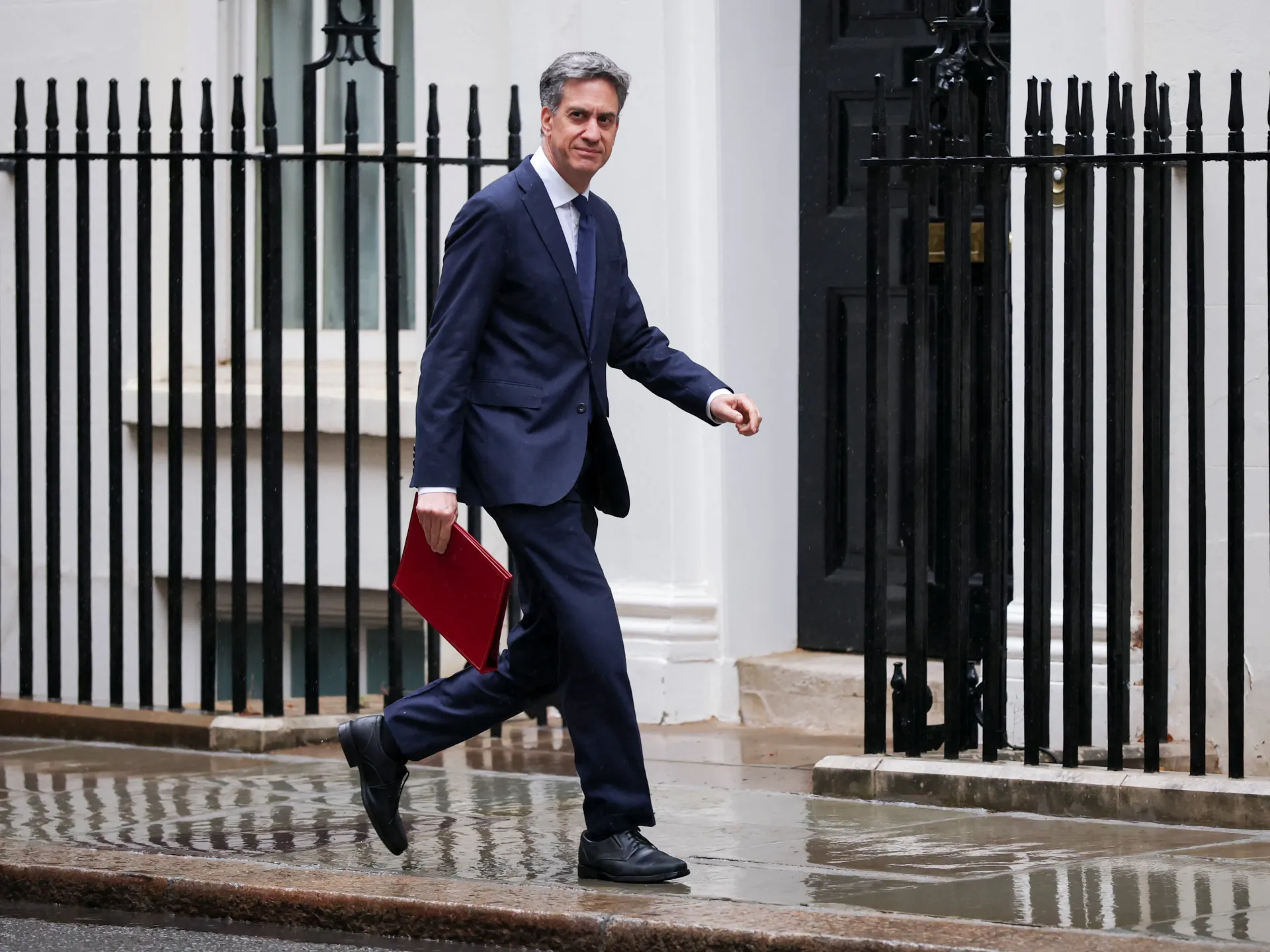 Man in dark suit carrying a red folder walks past black railings on a wet pavement.