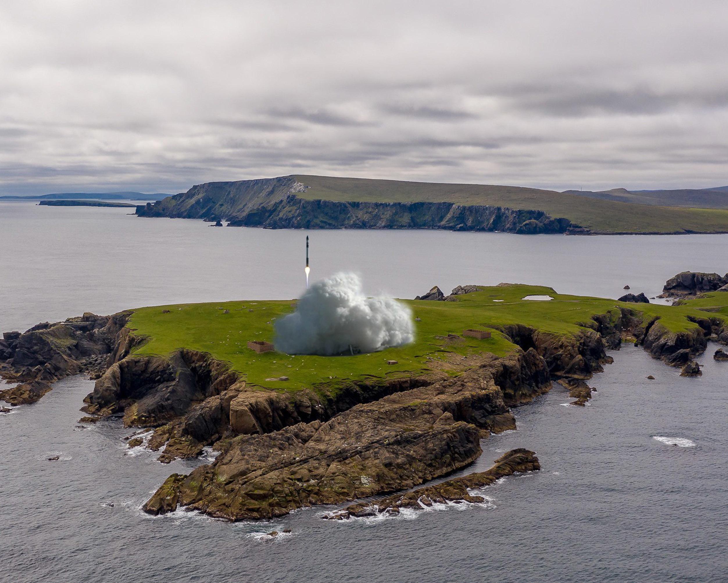 A cloudy sky over rocky green islands, with a plume of smoke rising near the shore.