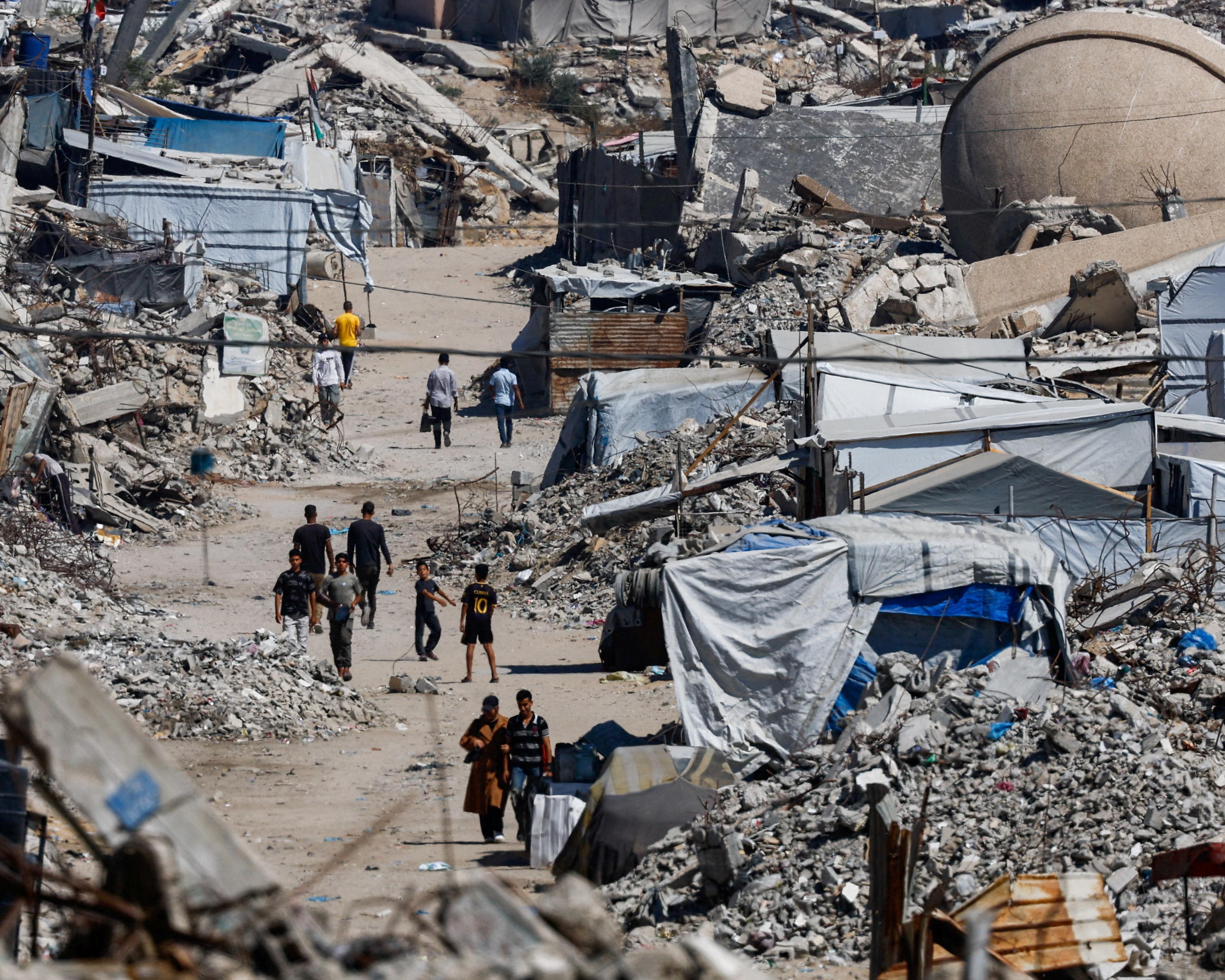People walk along a dusty path through extensive rubble and makeshift shelters.
