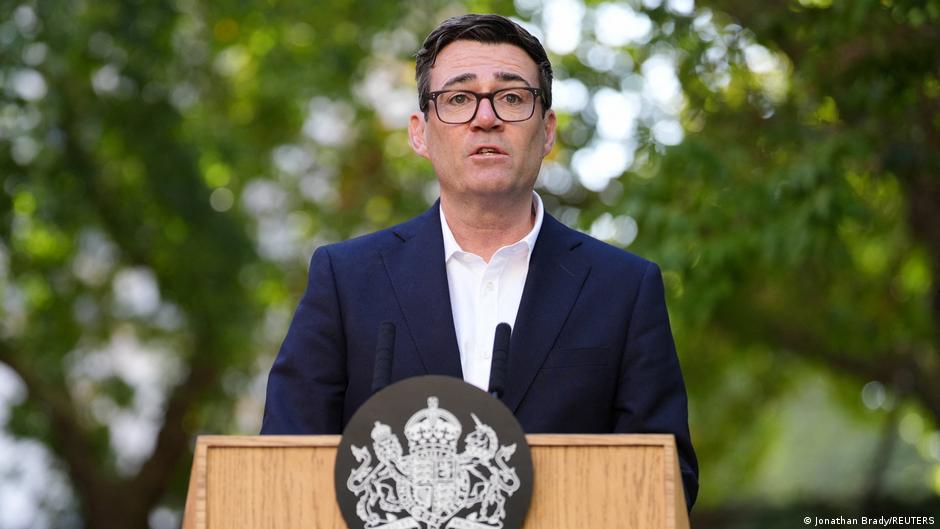 A suited man wearing glasses speaks at a government-emblem podium outdoors among trees.