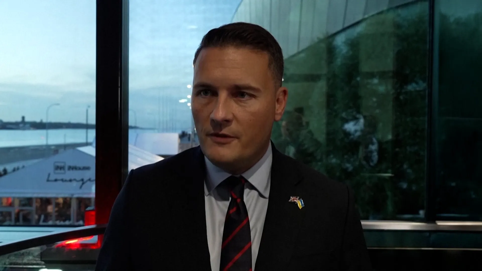 A suited man wearing small UK and Ukrainian flag pins speaks indoors beside large windows.