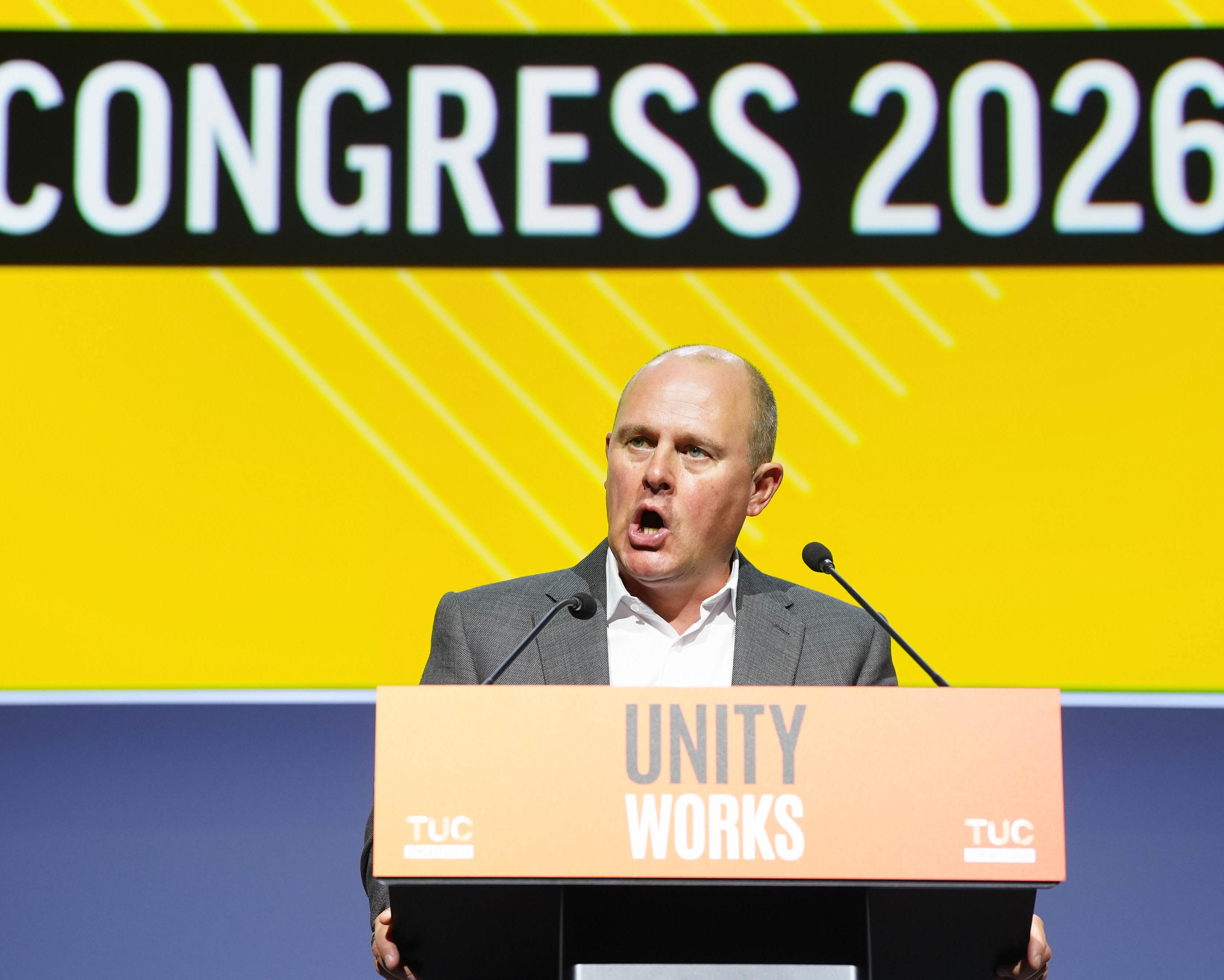 Man speaking at a podium beneath a Congress 2026 banner and Unity Works sign