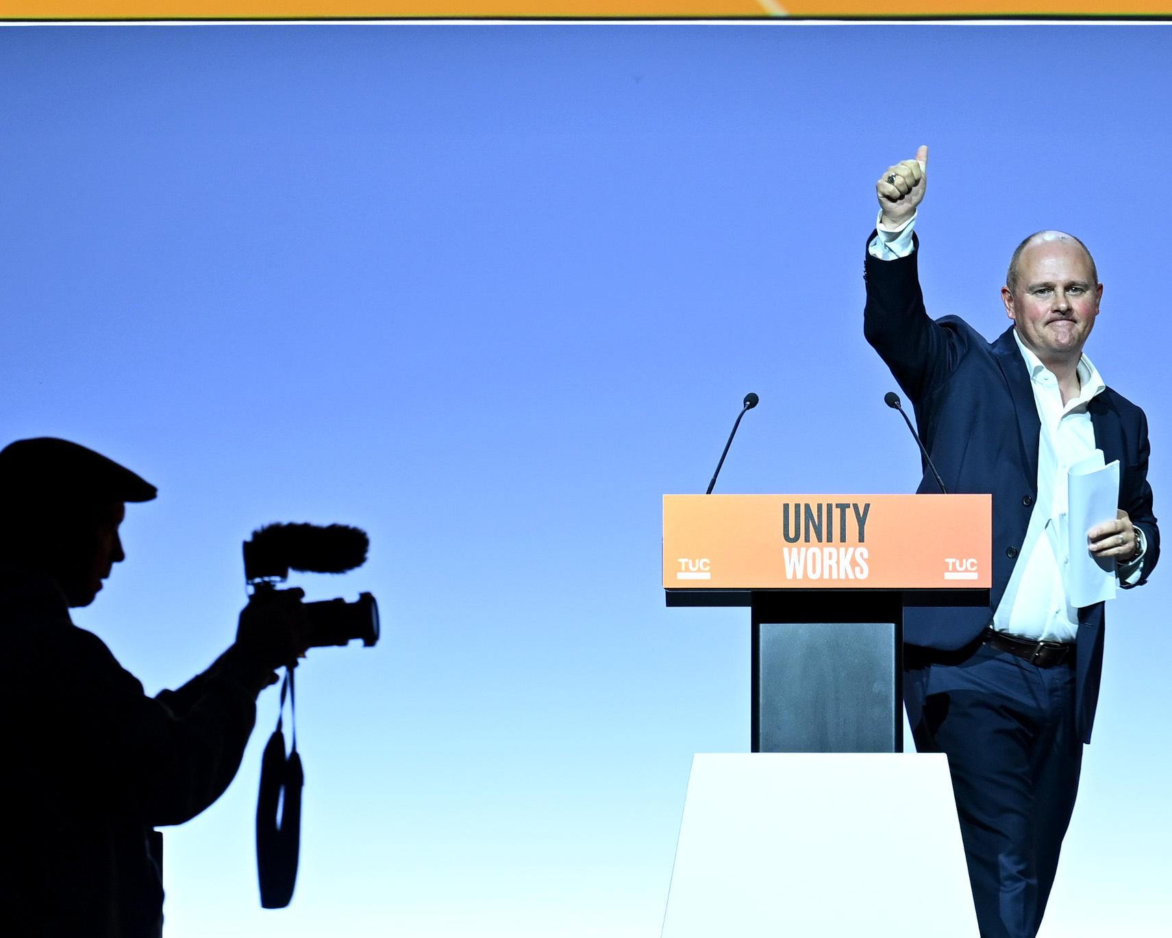 Speaker raises a fist beside a Unity Works podium as a silhouetted camera operator films from the foreground.