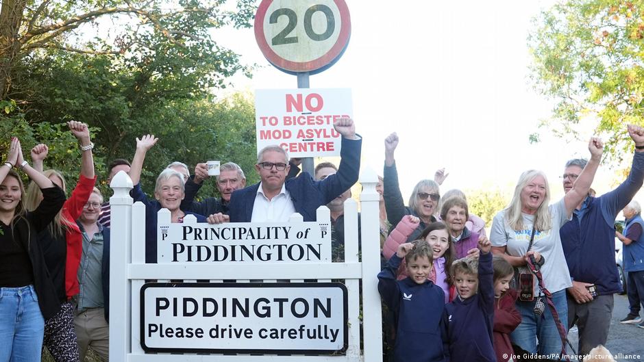 Group of villagers raising fists beside signs reading Principality of Piddington and Piddington Please drive carefully