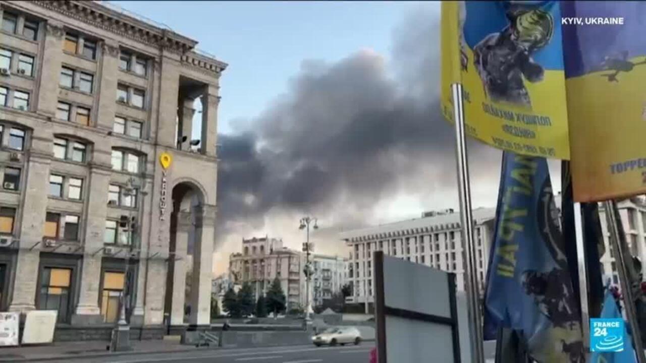 Dark smoke rises behind city buildings beside Ukrainian flags and a political billboard.