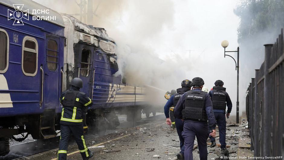 Emergency responders inspect a smoke-filled, damaged blue passenger train beside railway tracks.
