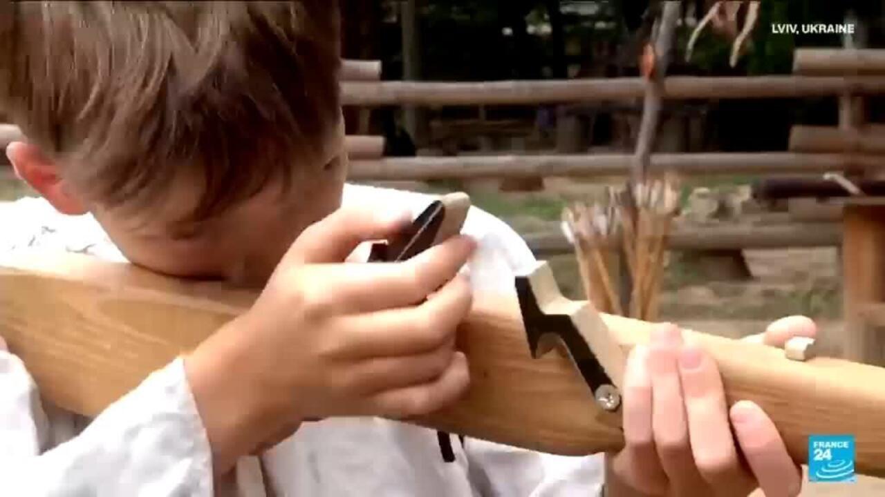 Child in white clothing holds a wooden bow-like device outdoors while gripping its mechanism.