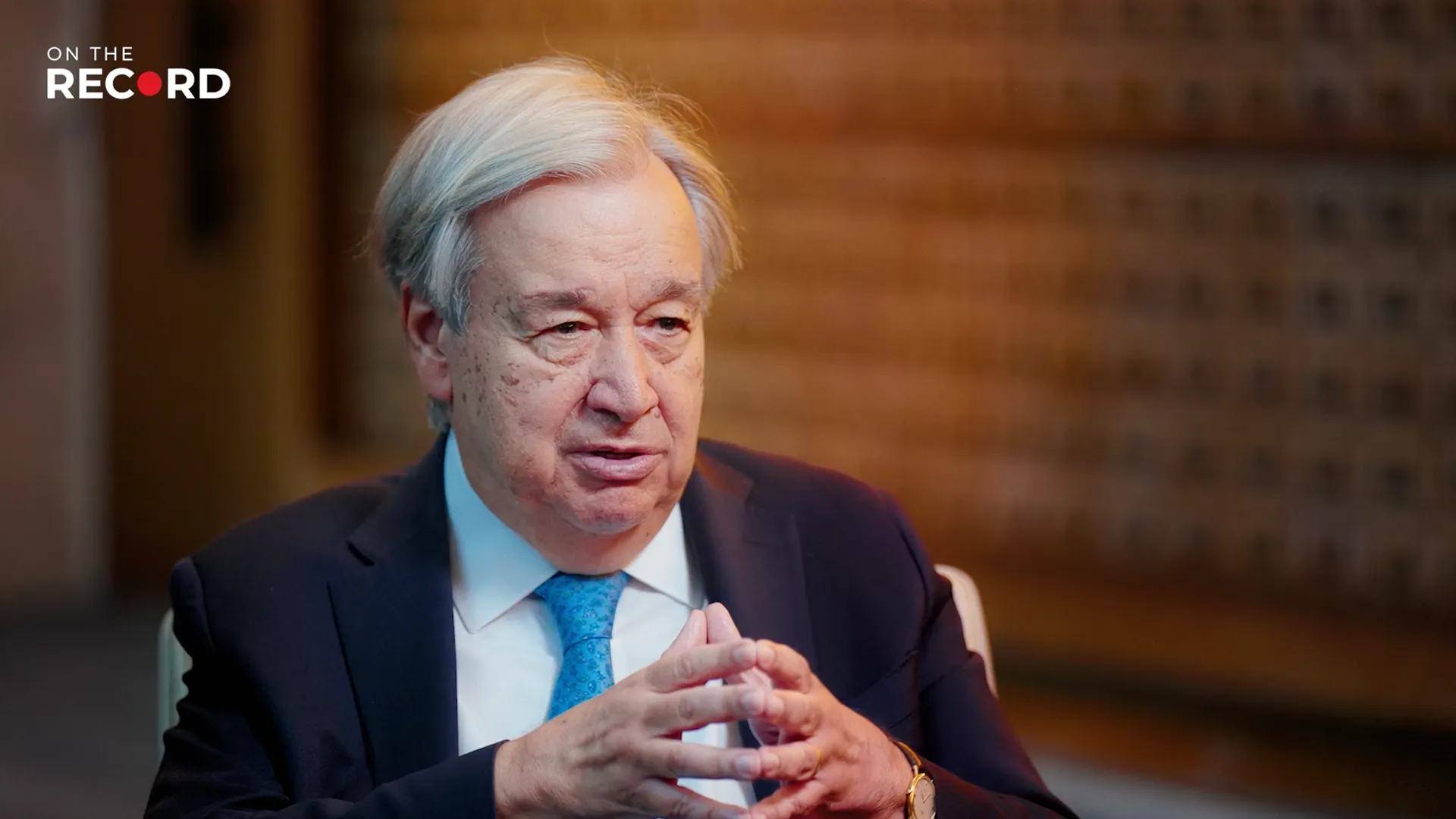 Gray-haired man in a dark suit and blue tie speaking during an interview.