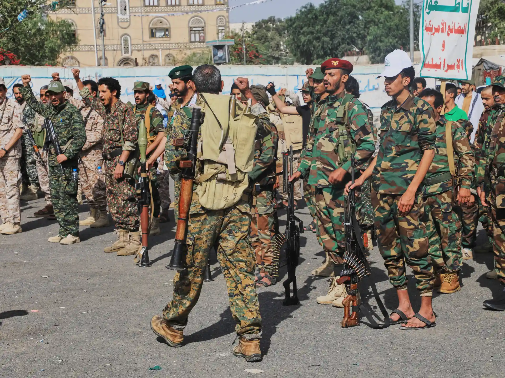 Armed men in camouflage stand in formation, several carrying rifles and ammunition belts during a street gathering.