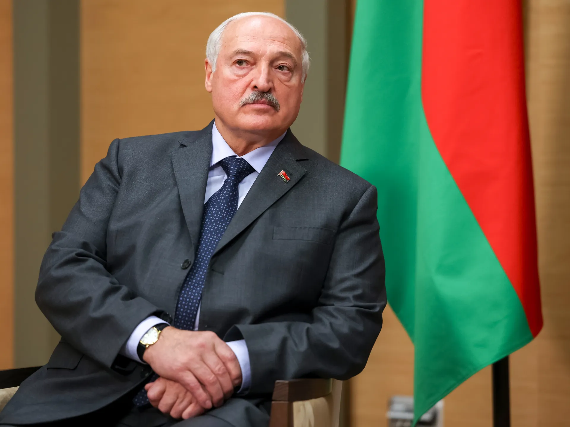 A suited older man sits with folded hands before a Belarusian flag.