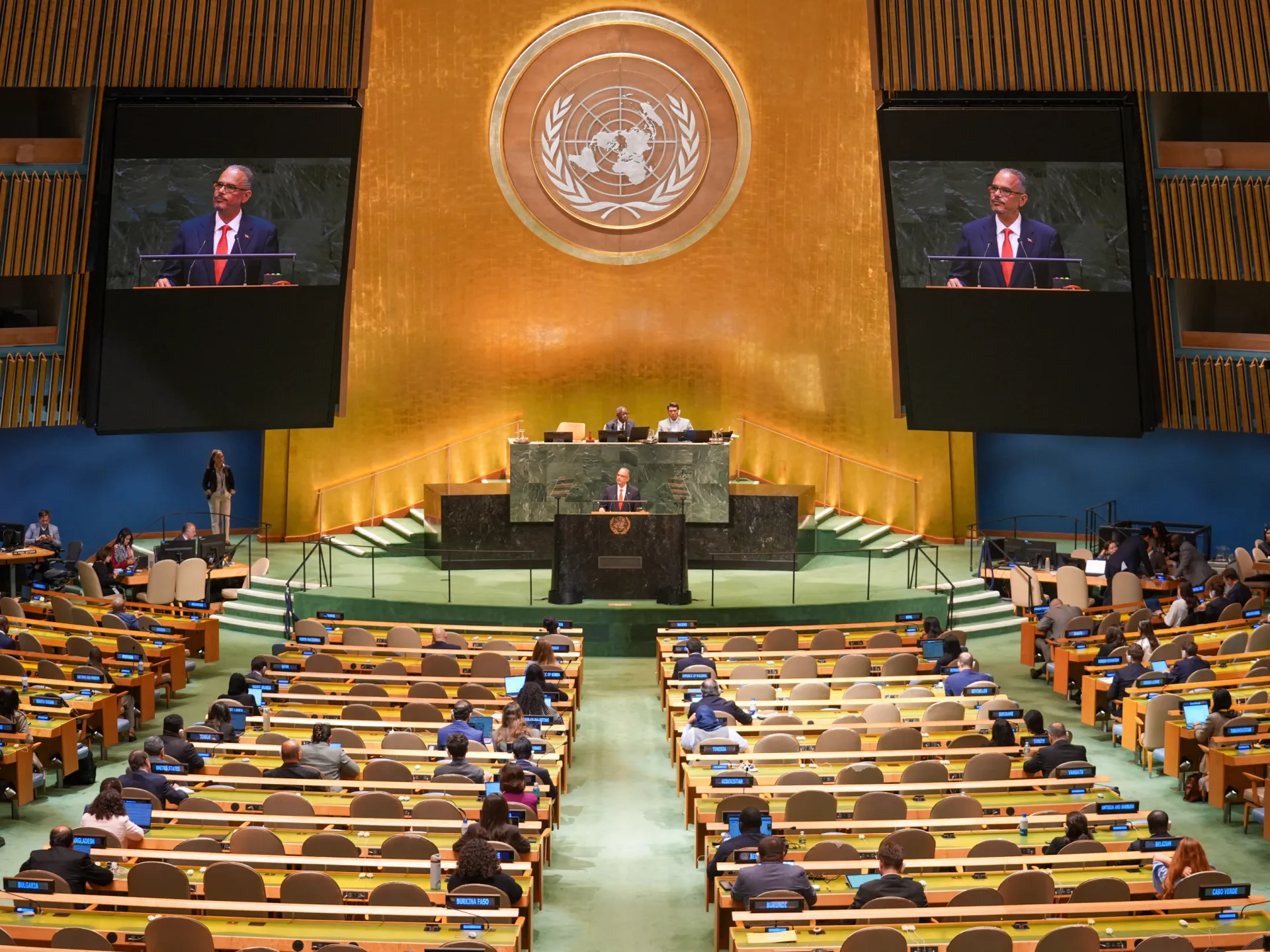 Delegates sit in the UN General Assembly hall as a speaker appears on two large screens.