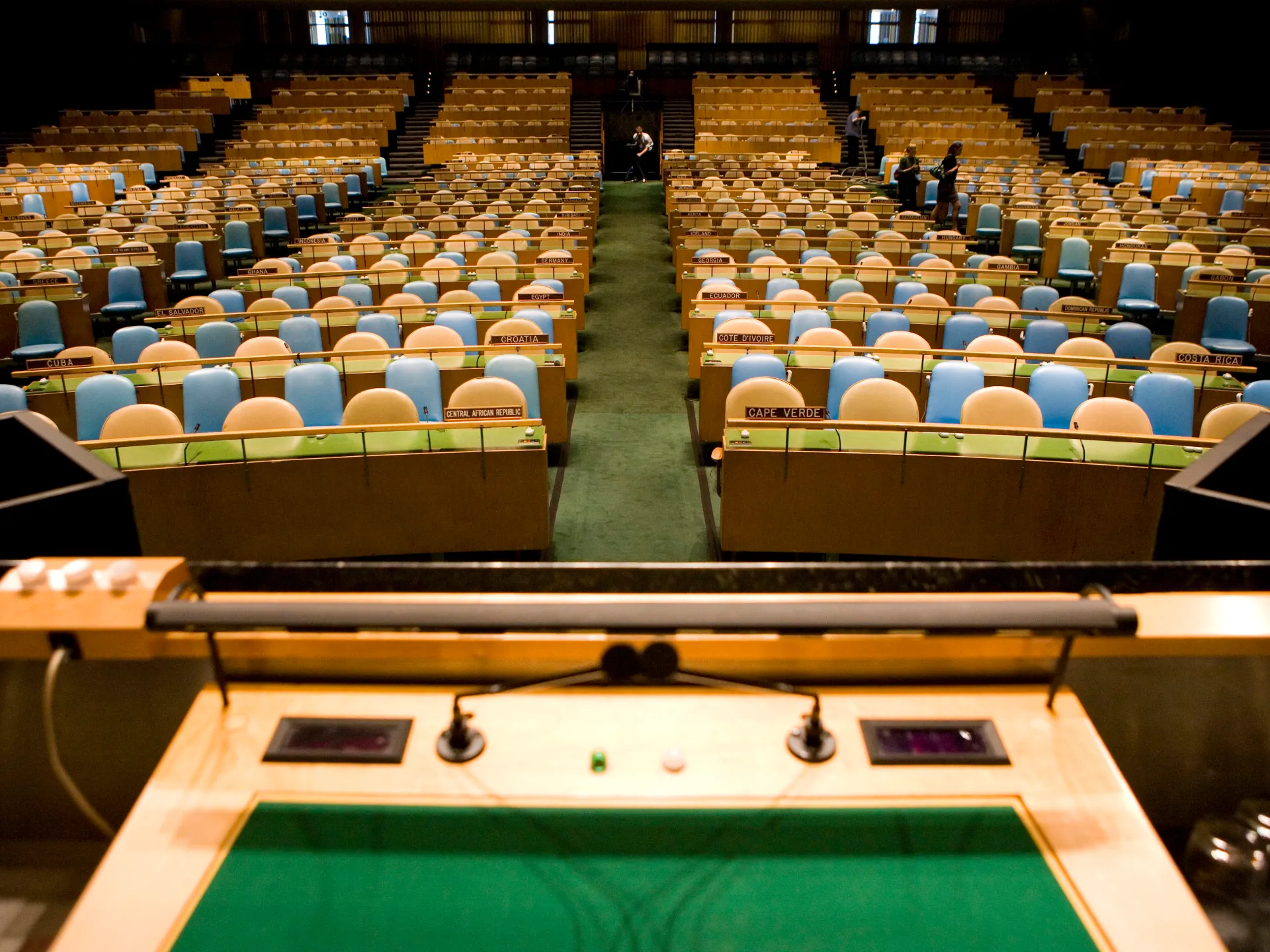 Empty international assembly hall with rows of blue and tan seats, country nameplates, and a central aisle.