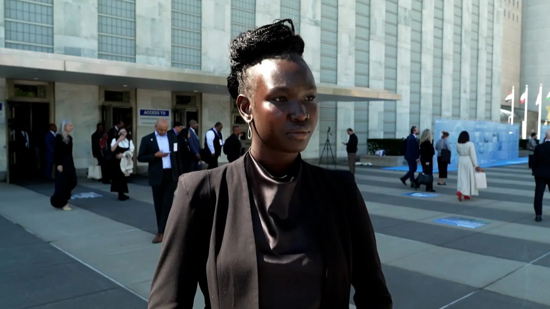 Woman in dark suit standing outside a large building with people gathered behind her