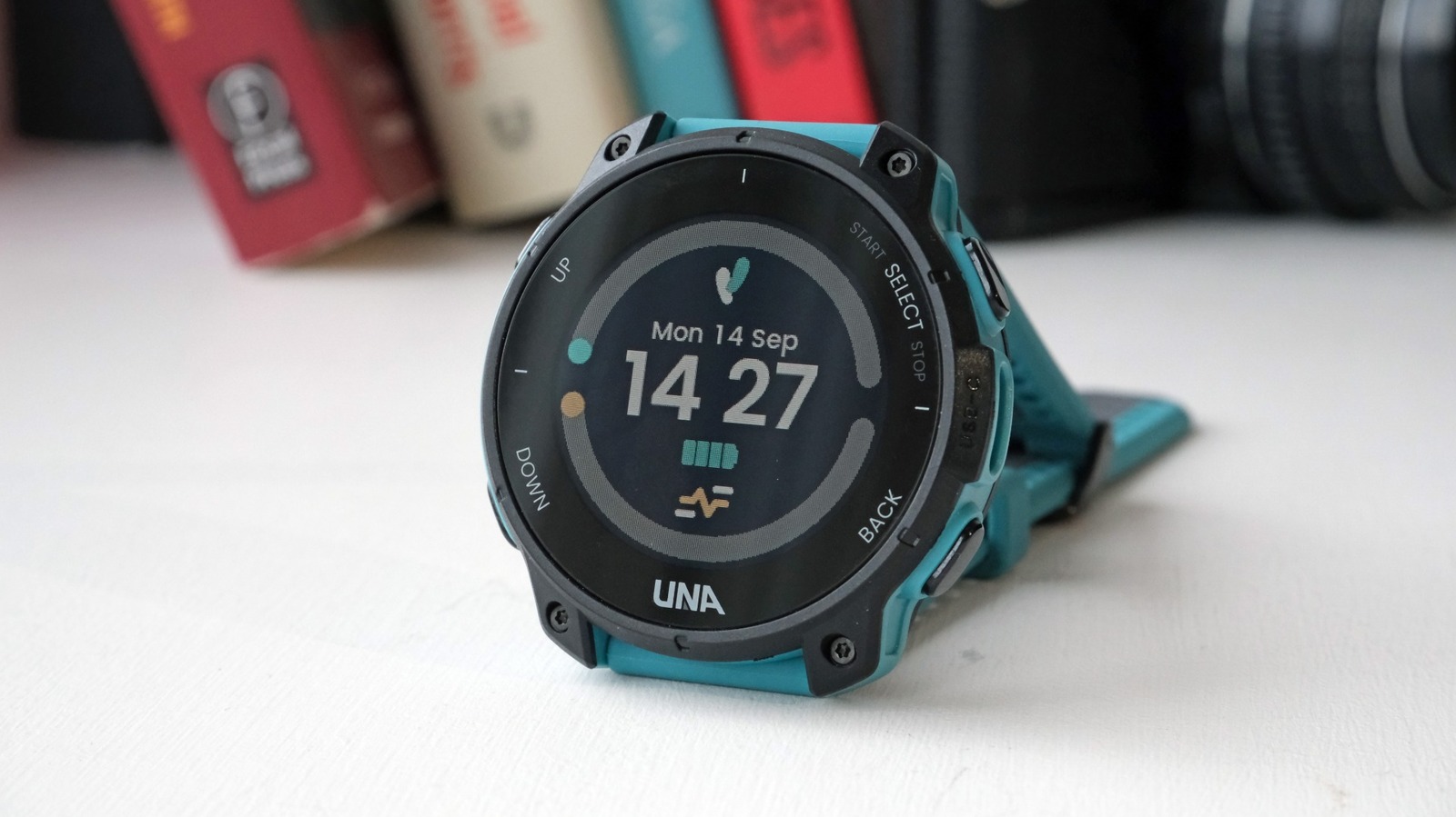 A teal and black rugged GPS watch displays the time on a white tabletop, with books and a camera lens behind it.