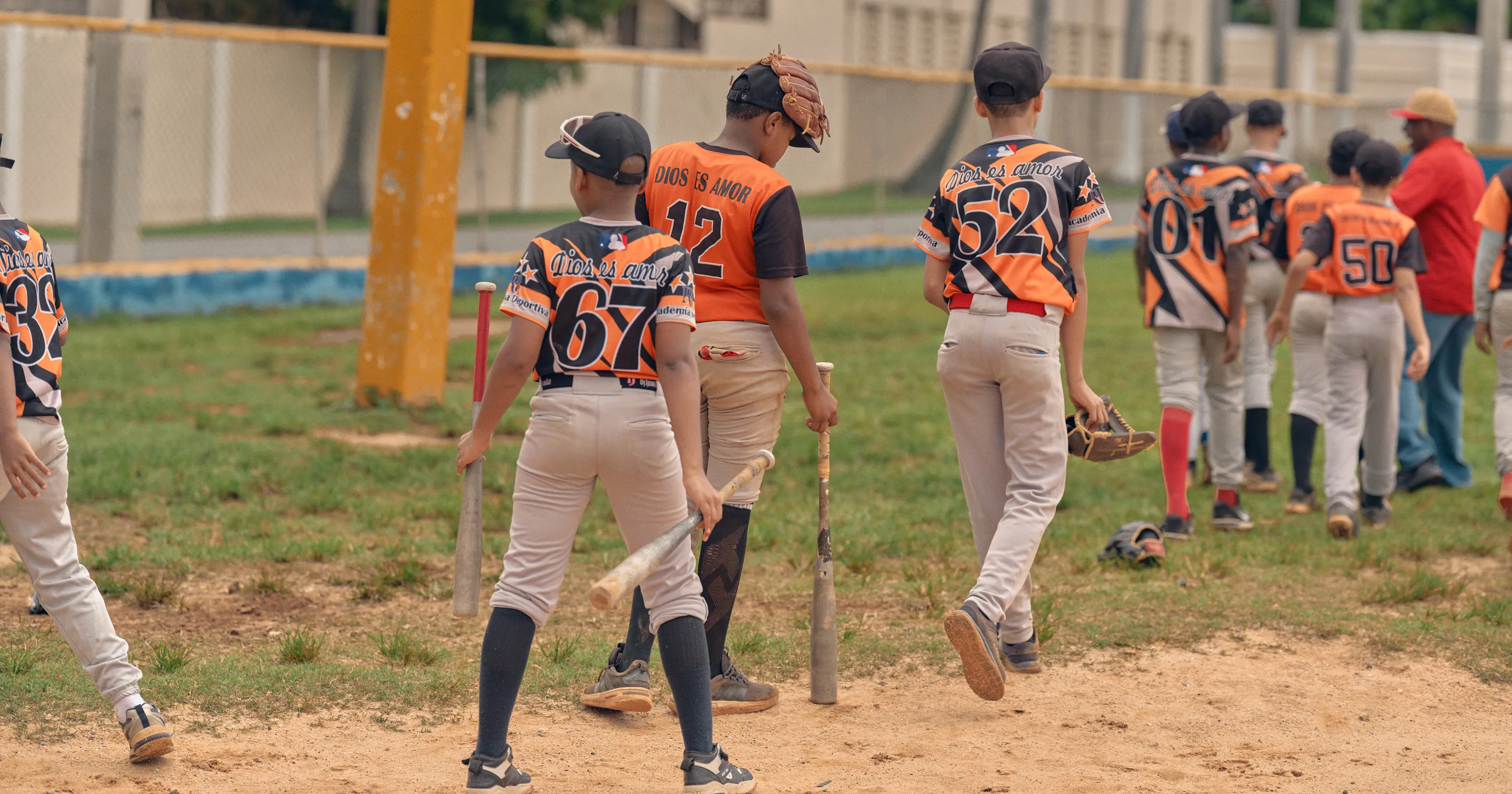 Young baseball players in orange-and-black uniforms walk across a dirt field carrying bats and gloves.