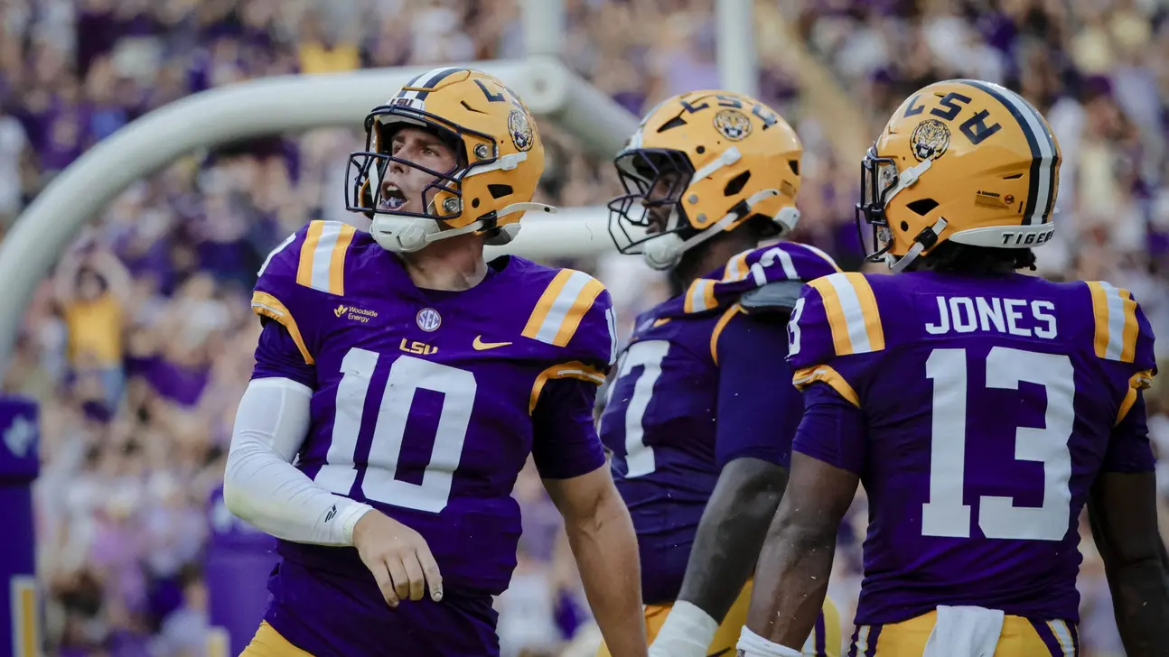 Three LSU football players in purple and gold uniforms stand together during a game.