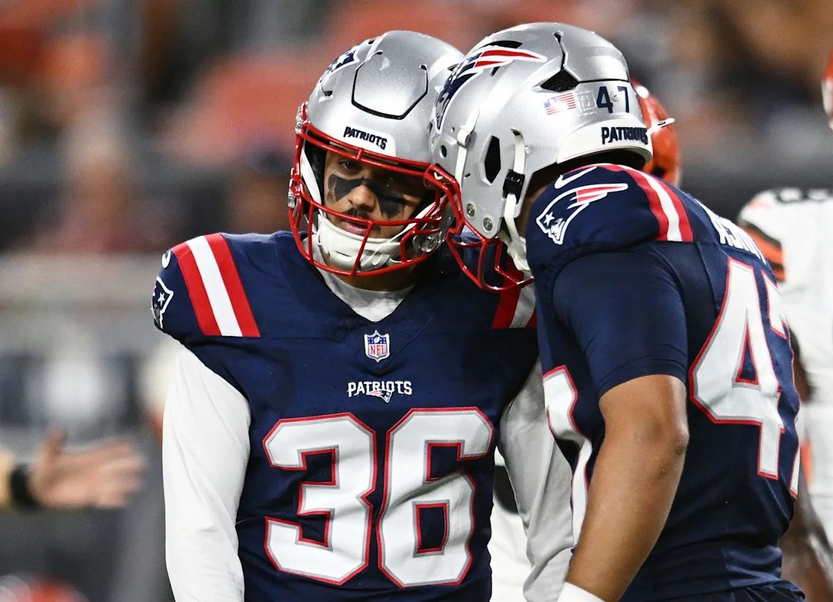 Two New England Patriots players in helmets and uniforms confer on the sideline during a game.