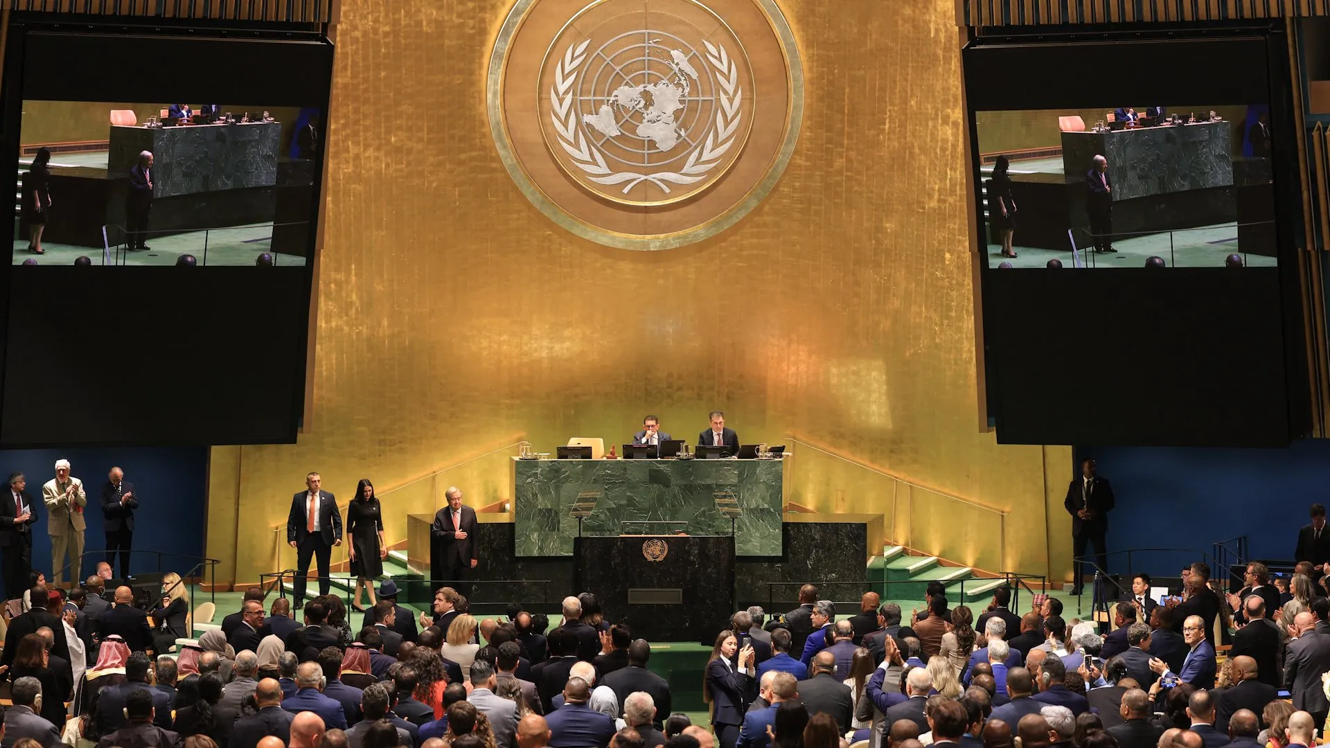 UN assembly hall with officials seated beneath the UN emblem as delegates applaud