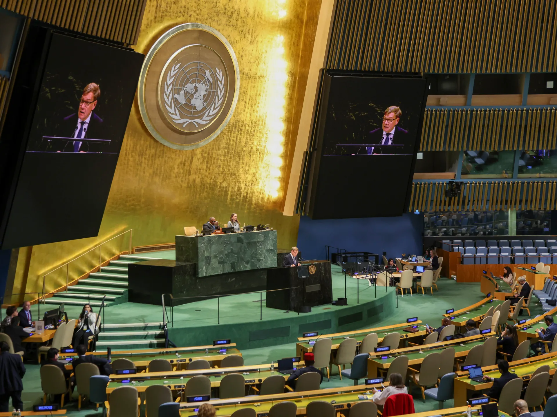 Wide view of an assembly hall with a speaker shown on two screens and delegates seated below.