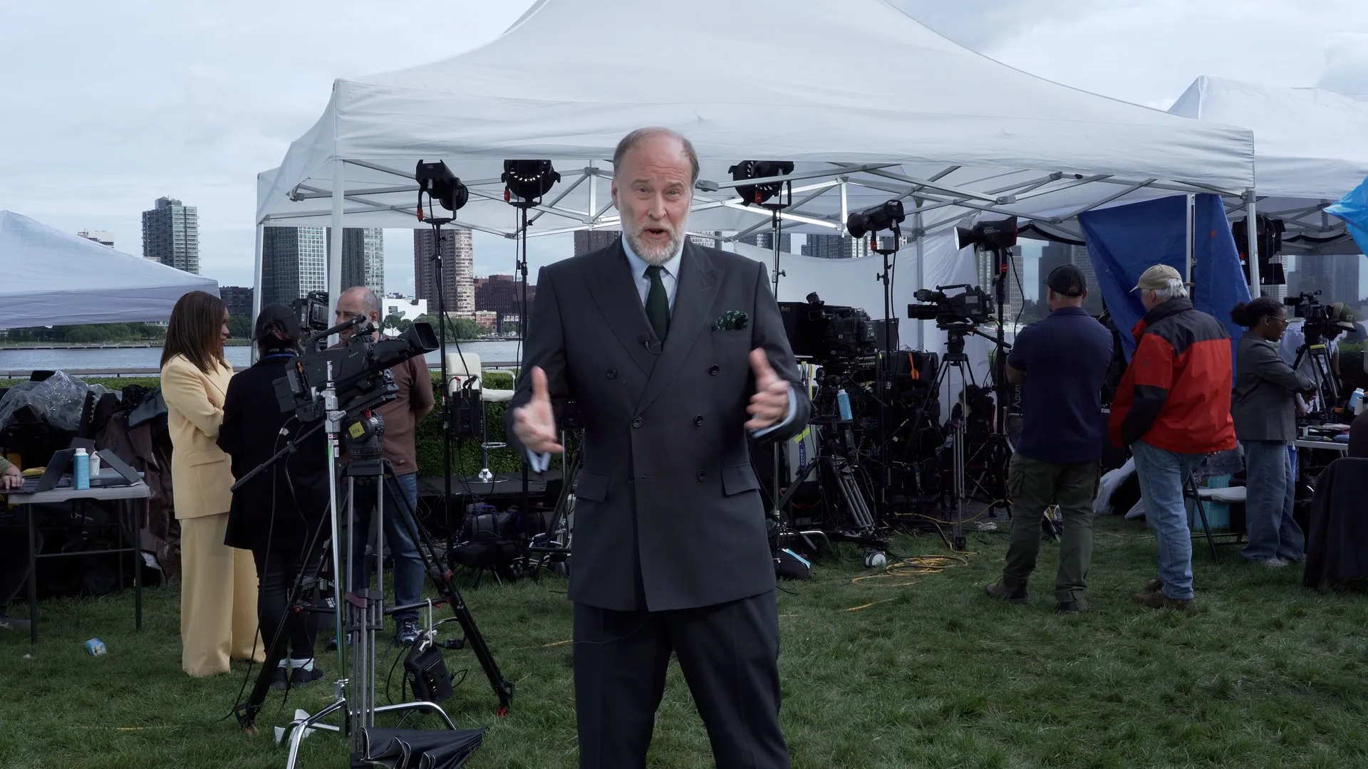 A suited man speaks to camera amid television crews and equipment beneath an outdoor canopy.