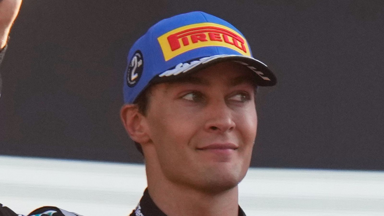 Race driver wearing a blue Pirelli cap looks sideways with a slight smile