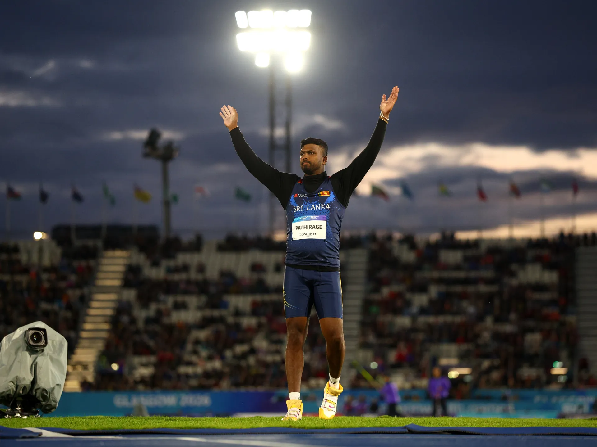 A Sri Lankan javelin athlete stands with both arms raised on the field as stadium lights glow behind him.