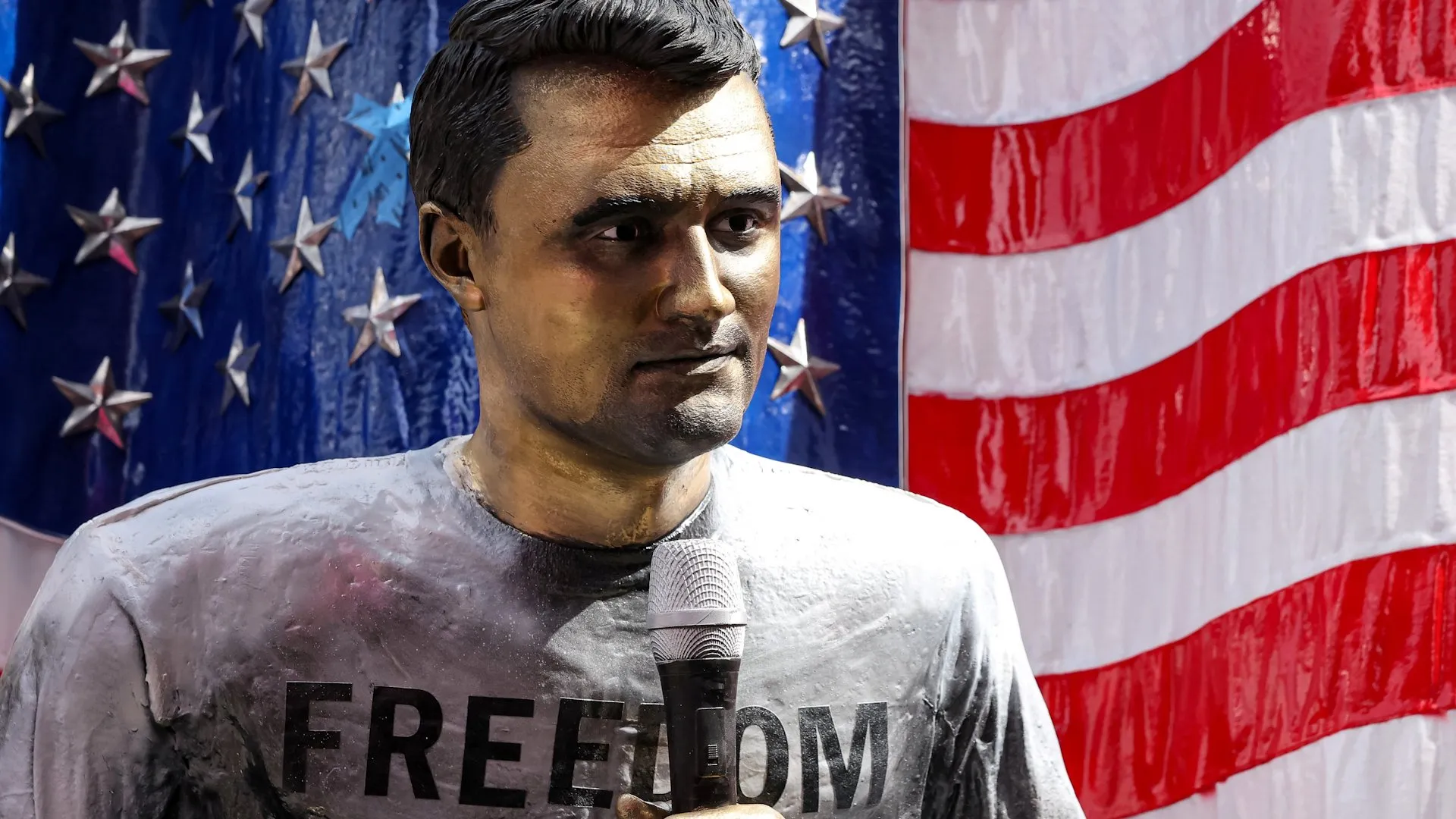 Statue of a man holding a microphone before American flag imagery, wearing a shirt marked “FREEDOM.”