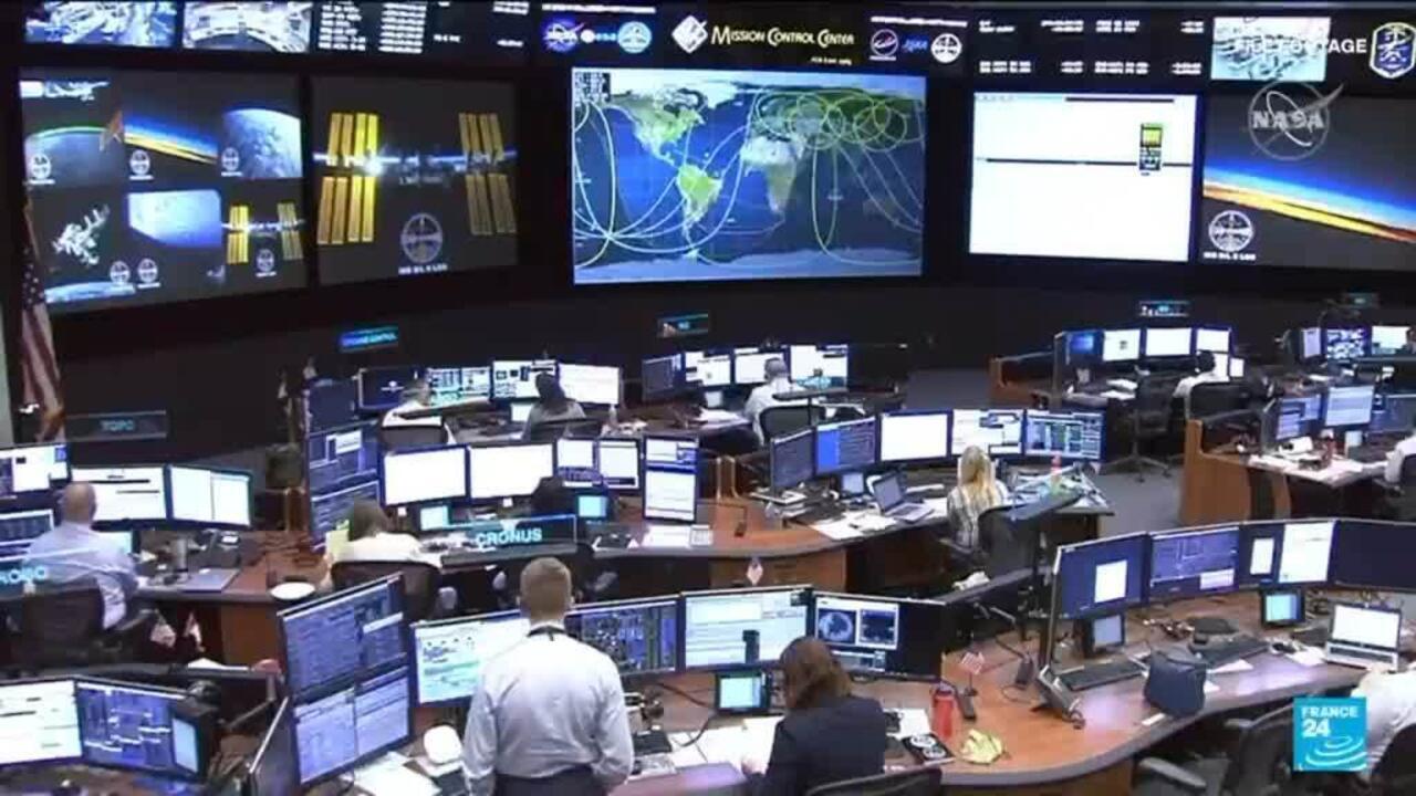 Mission control room filled with operators, computer consoles, and large displays showing space-related graphics.