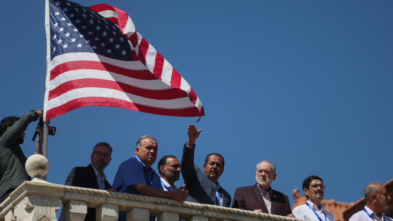 U.S. flag waves above a group of people gathered on a stone balcony under a clear blue sky.