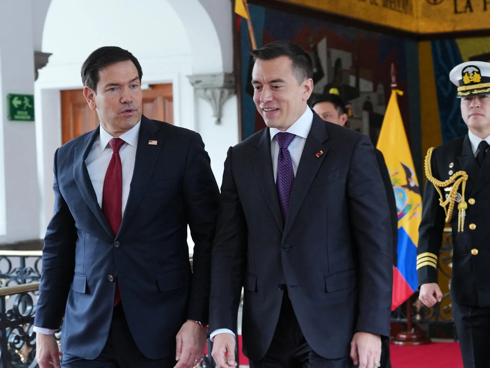 Two suited officials walk beside a uniformed naval officer near an Ecuadorian flag.