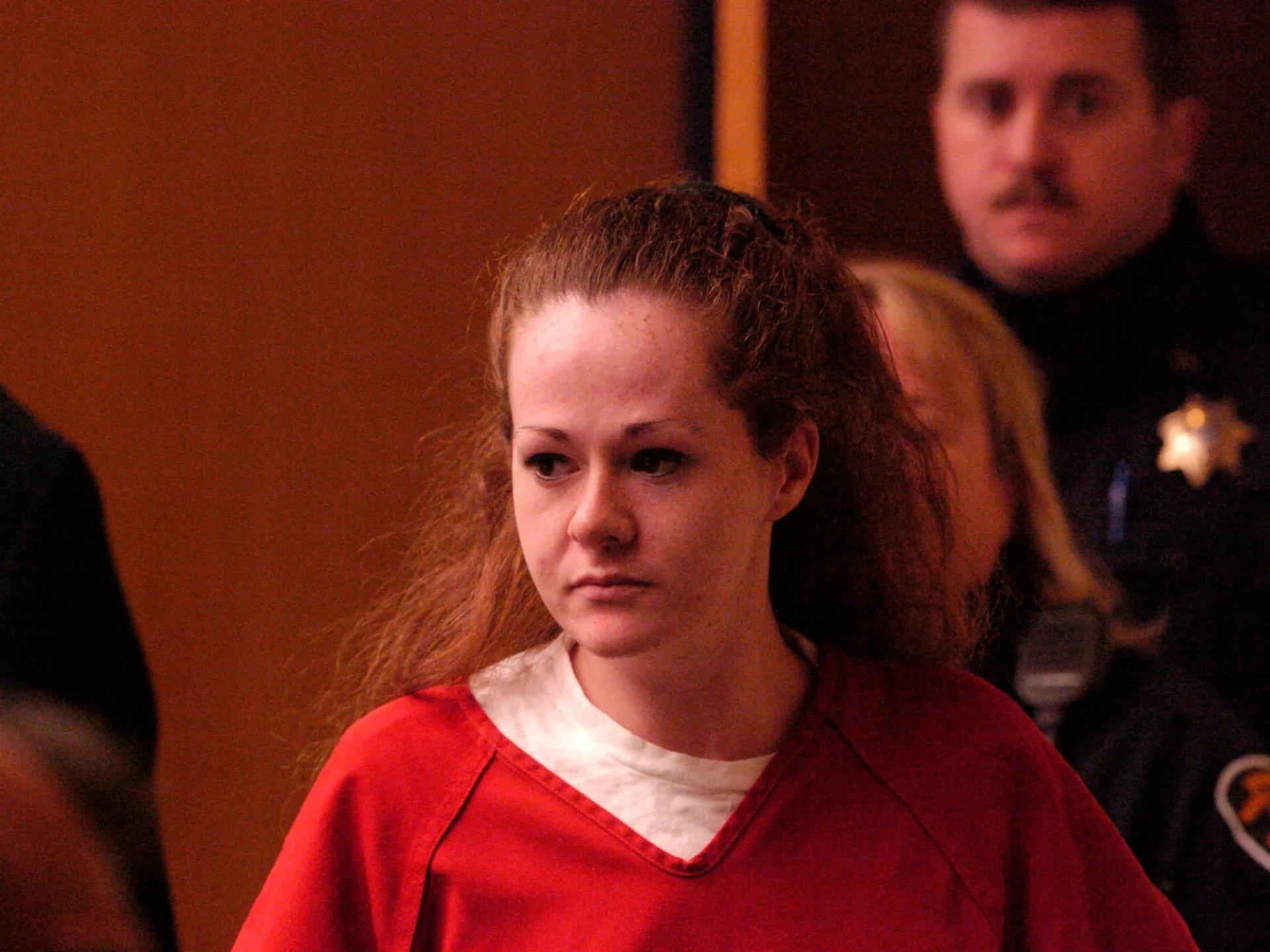 Woman in red prison clothing sits in a courtroom, with a uniformed officer behind her.