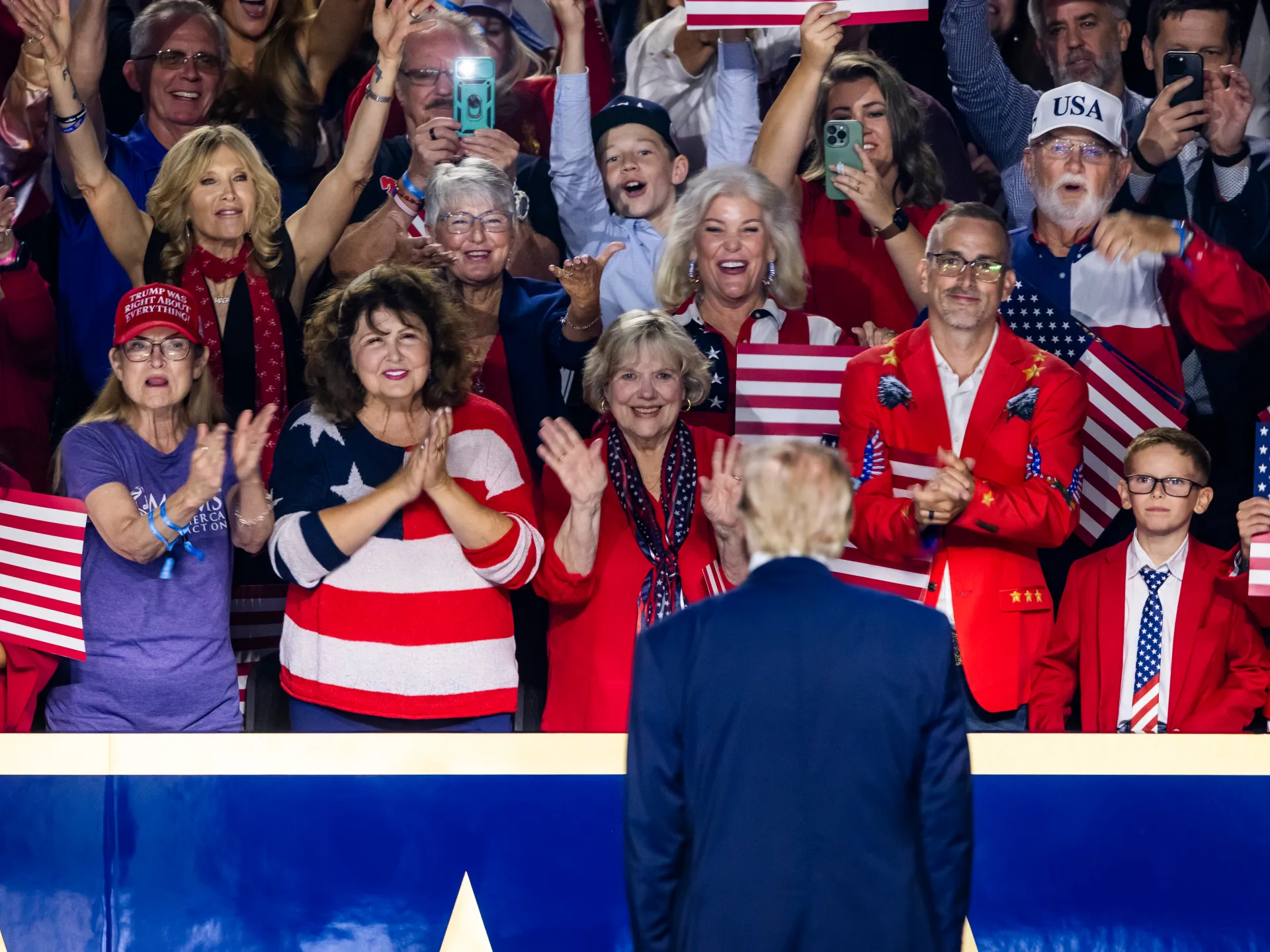 Crowd of supporters waves flags and applauds a speaker seen from behind at a political event.