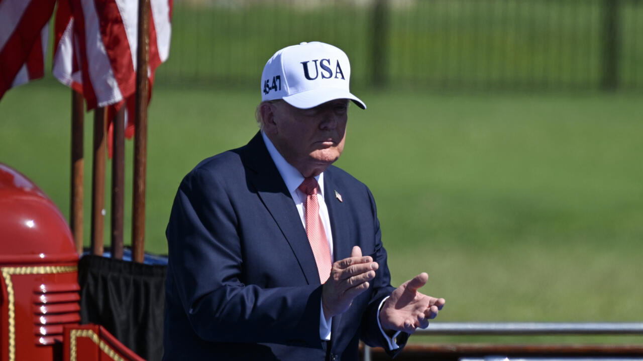 Man in dark suit and USA cap claps beside flags at an outdoor event
