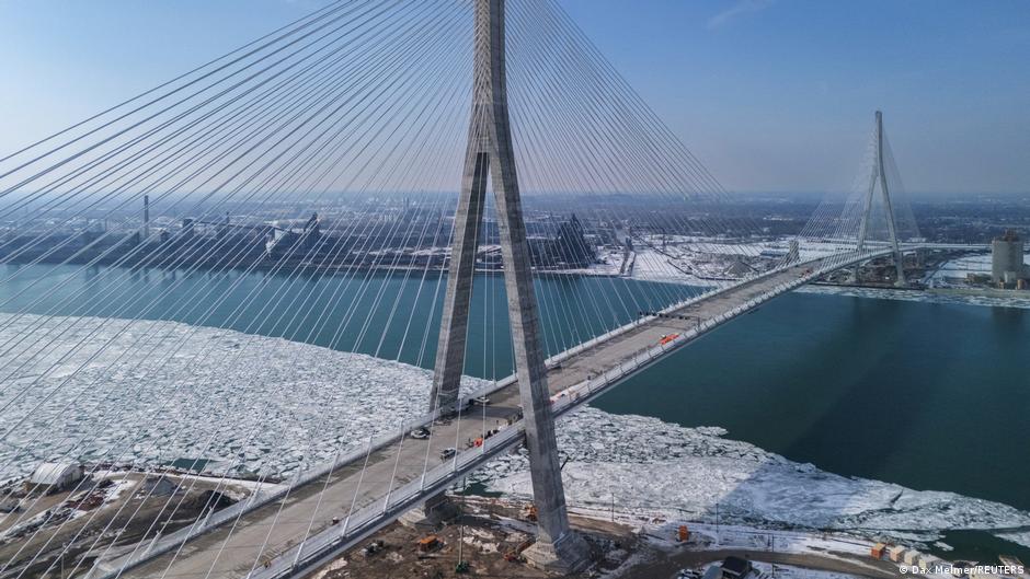 A cable-stayed bridge crosses ice-covered water beneath a hazy blue sky.